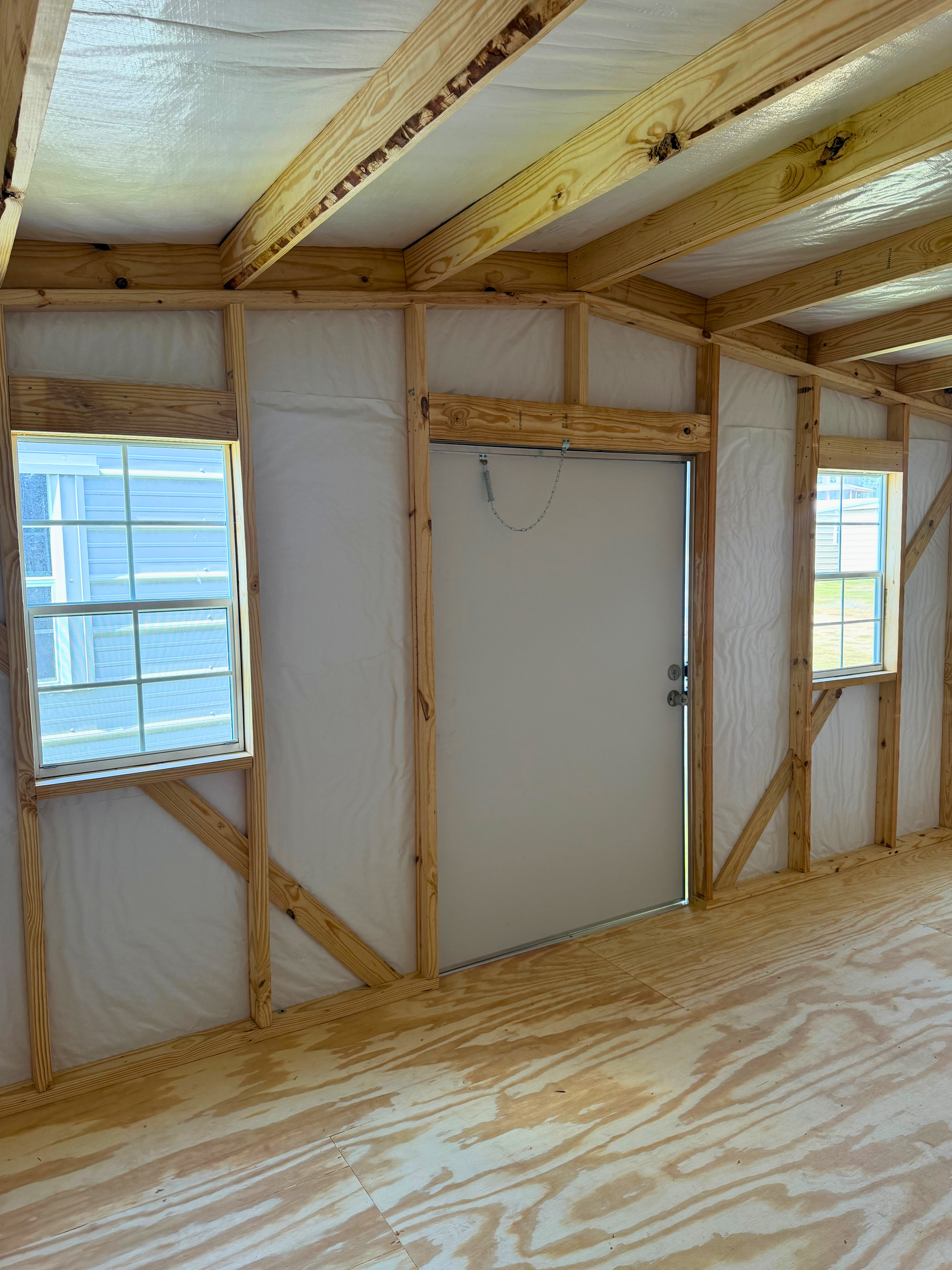 Interior of an unfinished room with exposed wood framing, windows, and a white door.