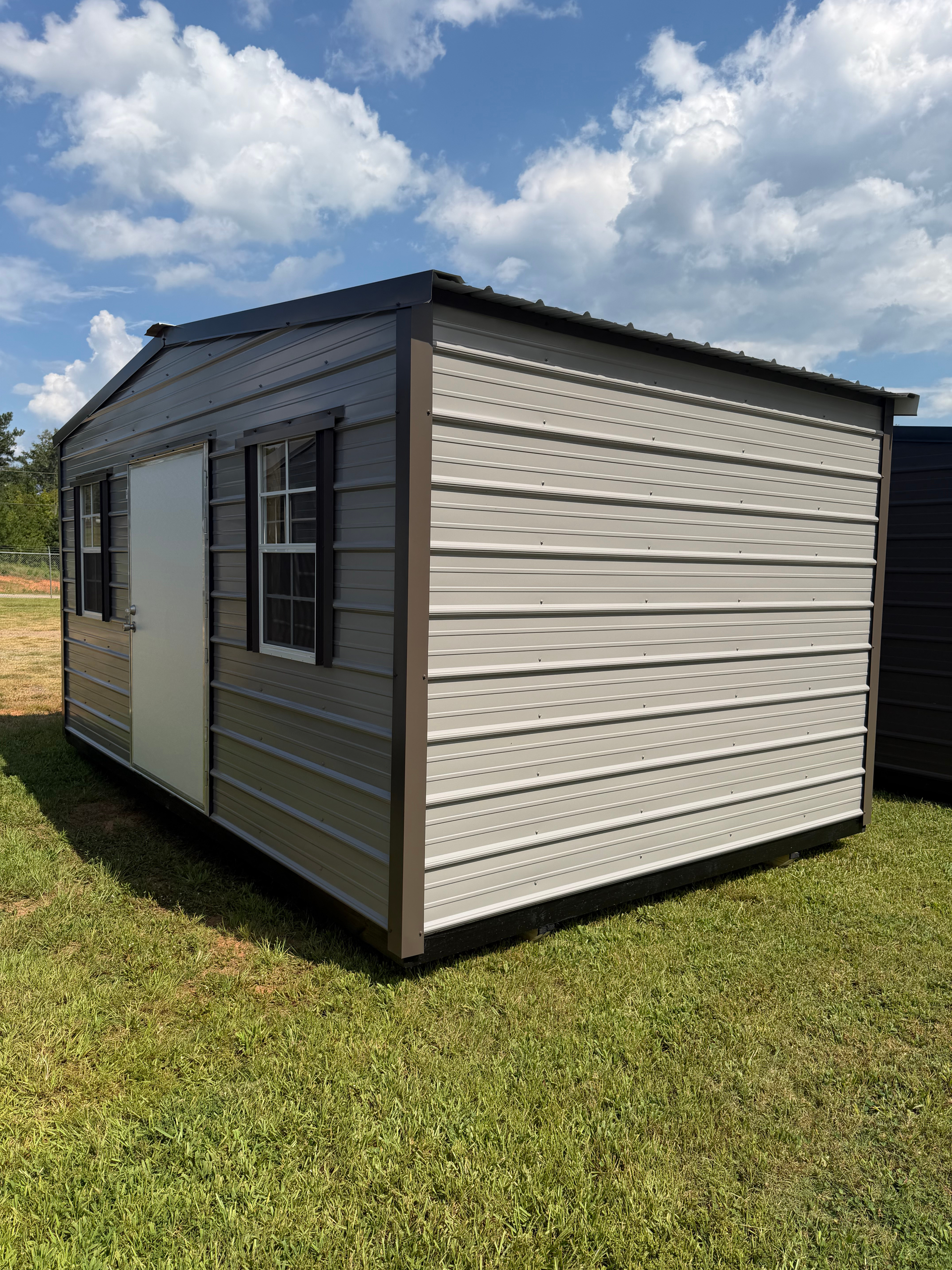 Small beige shed with black roof and trim on a grassy lawn under a partly cloudy sky