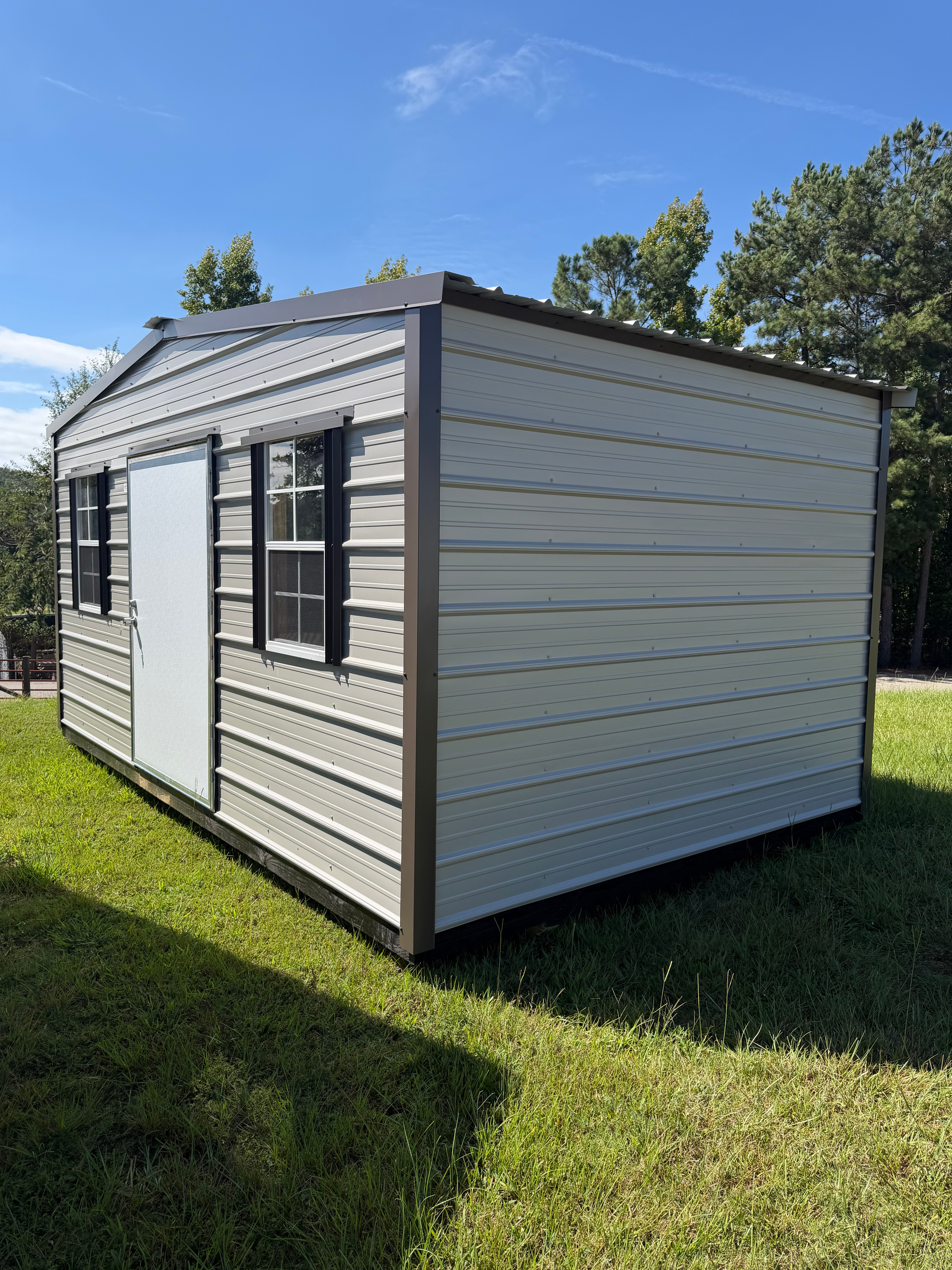 Small light-gray portable shed with windows on a grassy yard under a blue sky