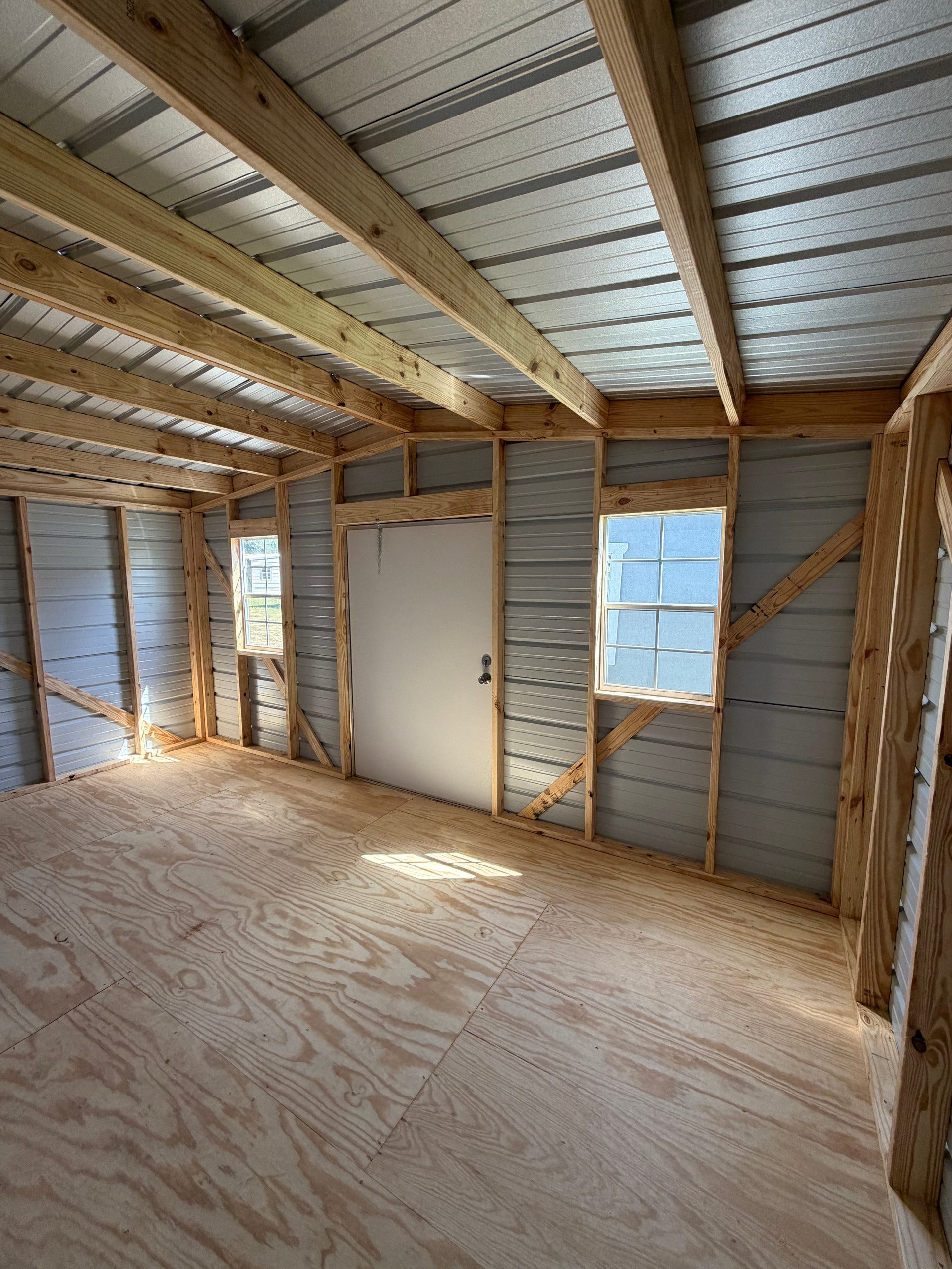 Empty unfinished room with exposed wood framing, plywood floor, and small window under construction