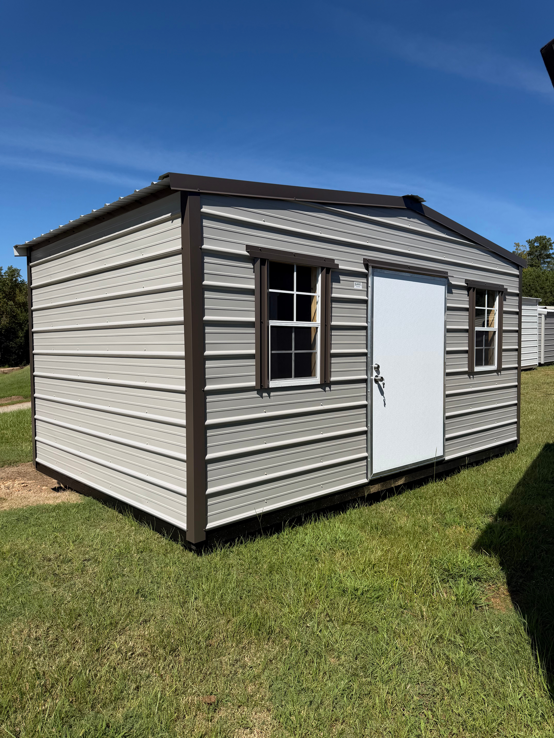 Small gray shed with dark trim, a white door, and windows, standing on grass under a blue sky.