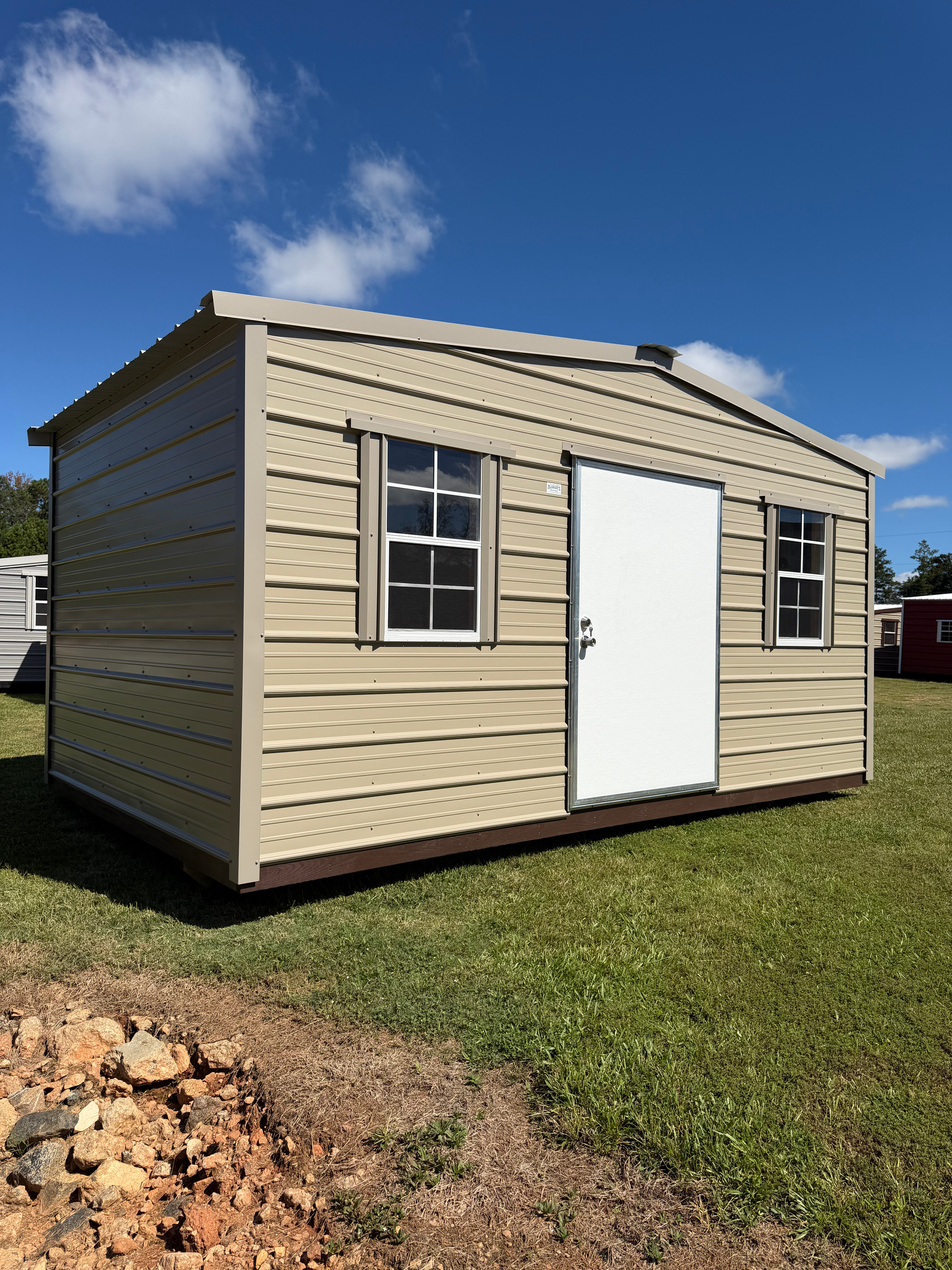 Small beige shed with a white door and windows, sitting on grass under a blue sky.