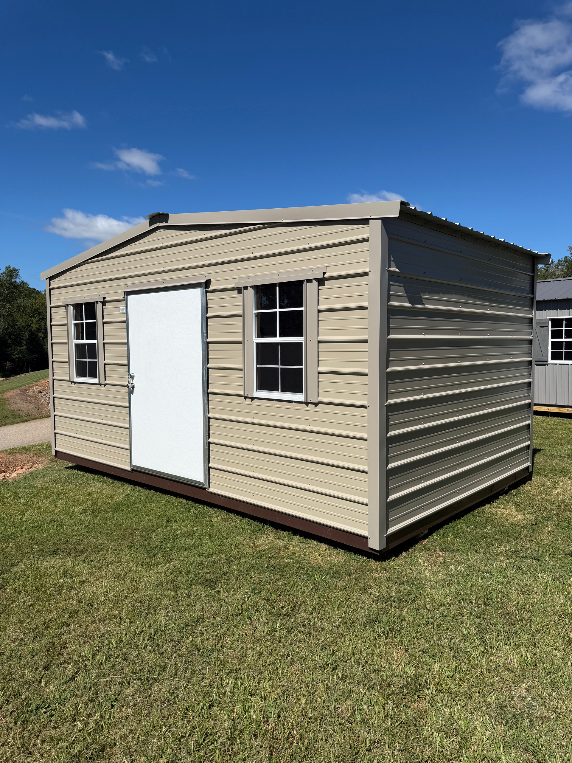 Small beige shed with white door and windows on a grassy lawn under a blue sky