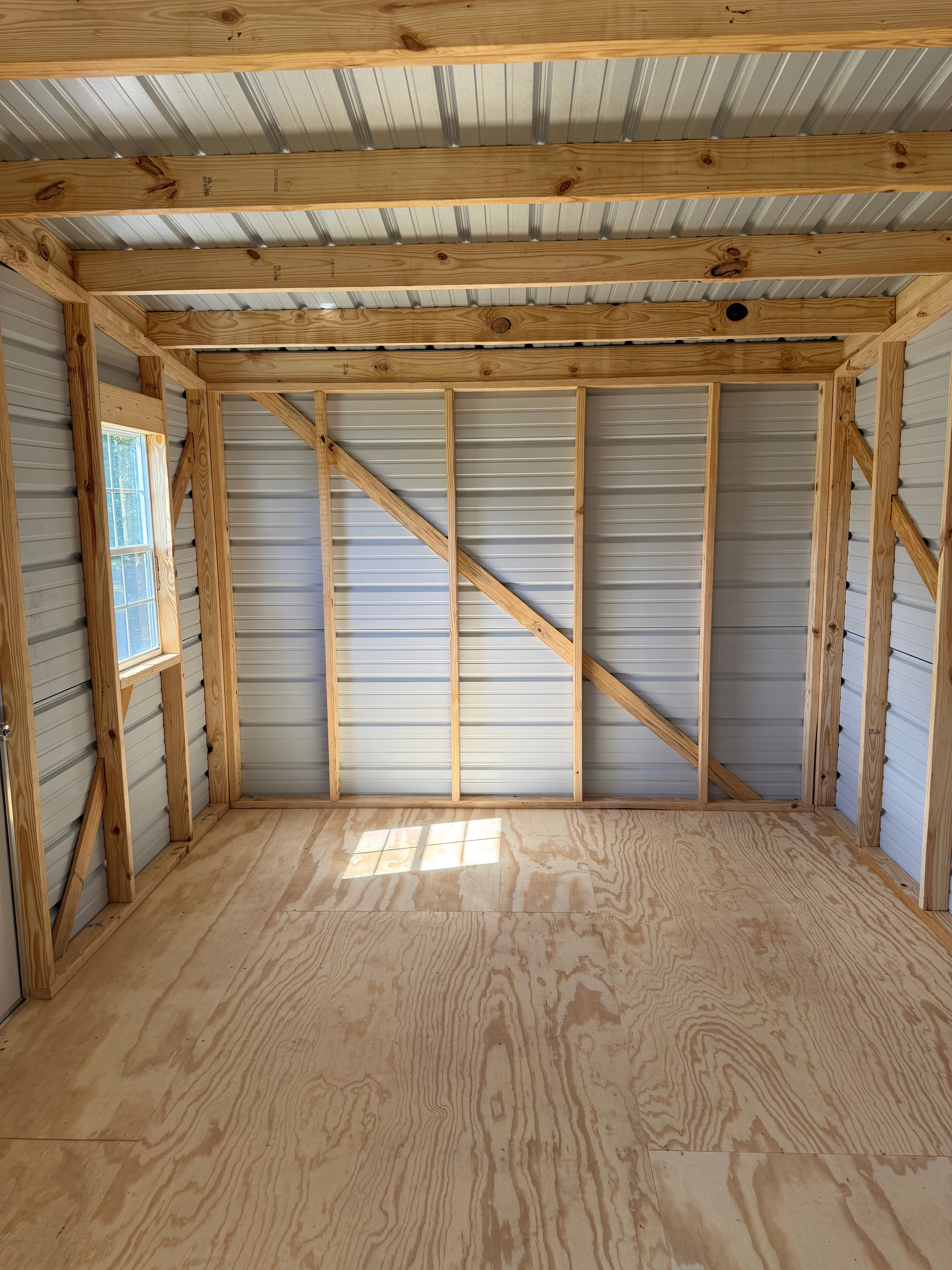 Empty unfinished wooden room with exposed studs, plywood floor, and light from a window opening.
