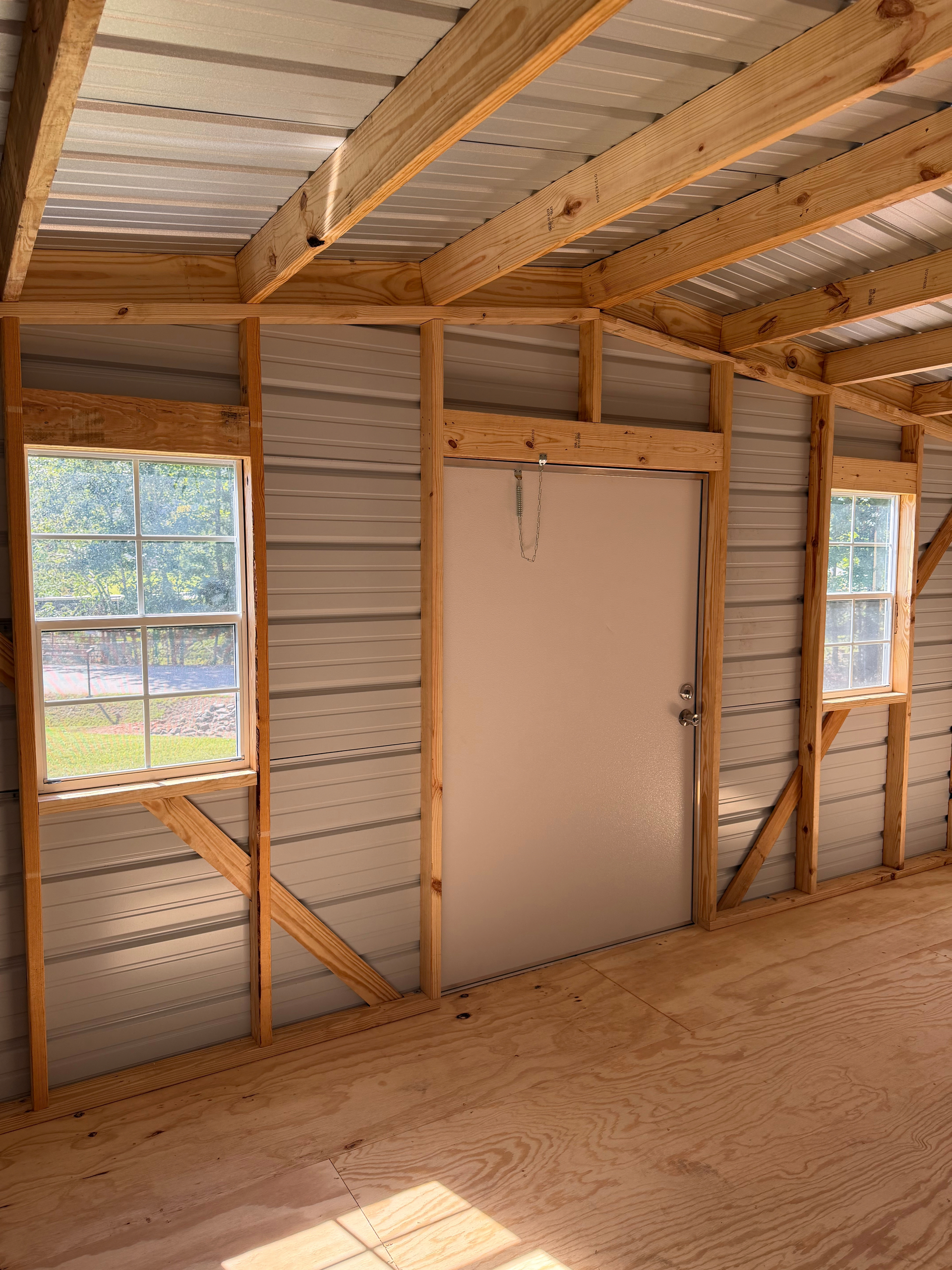 Unfinished wooden room interior with door and two windows, exposed framing, and sunlight on the floor.