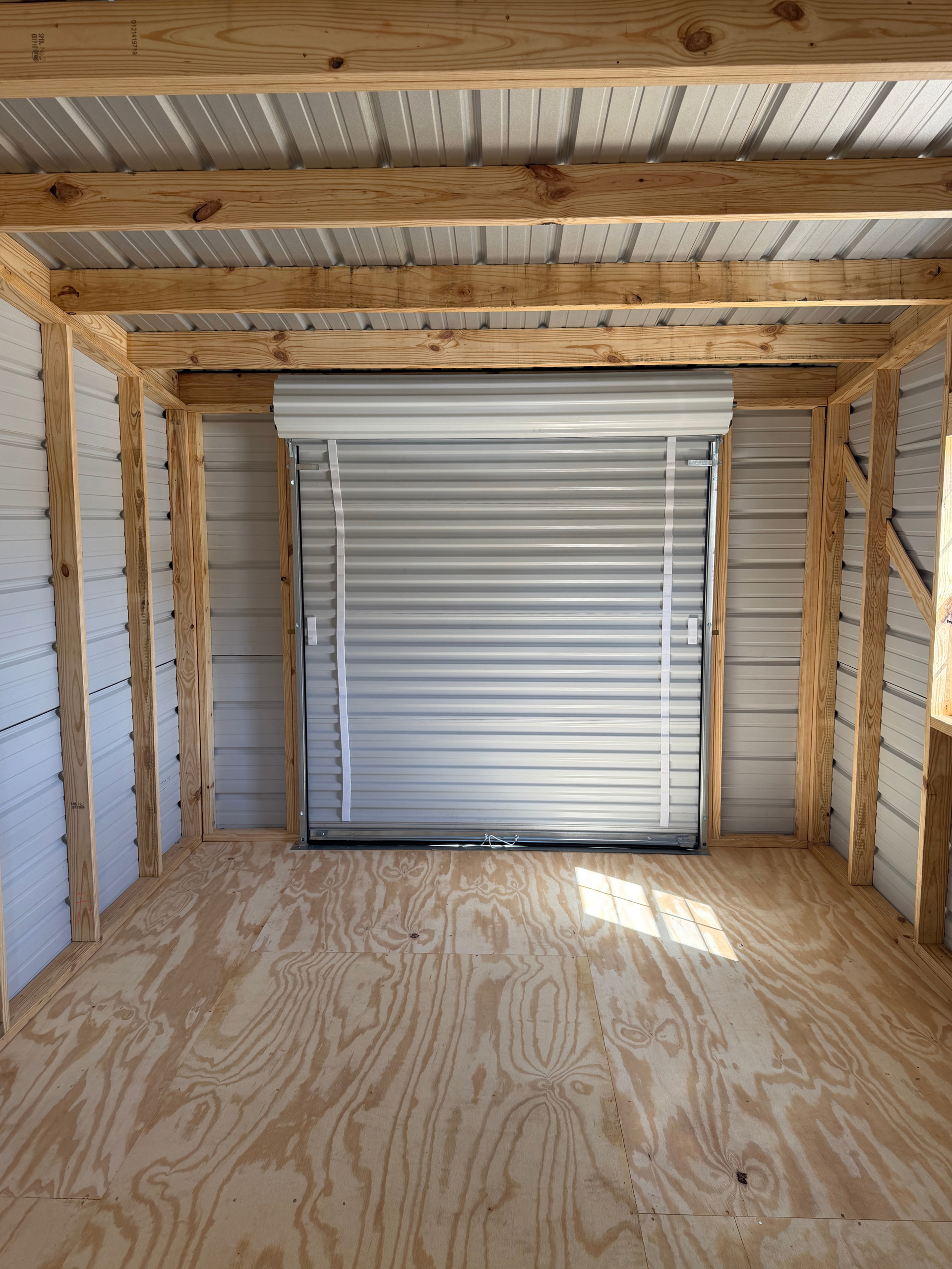 Empty wooden shed interior with a closed white roll-up door and plywood floor