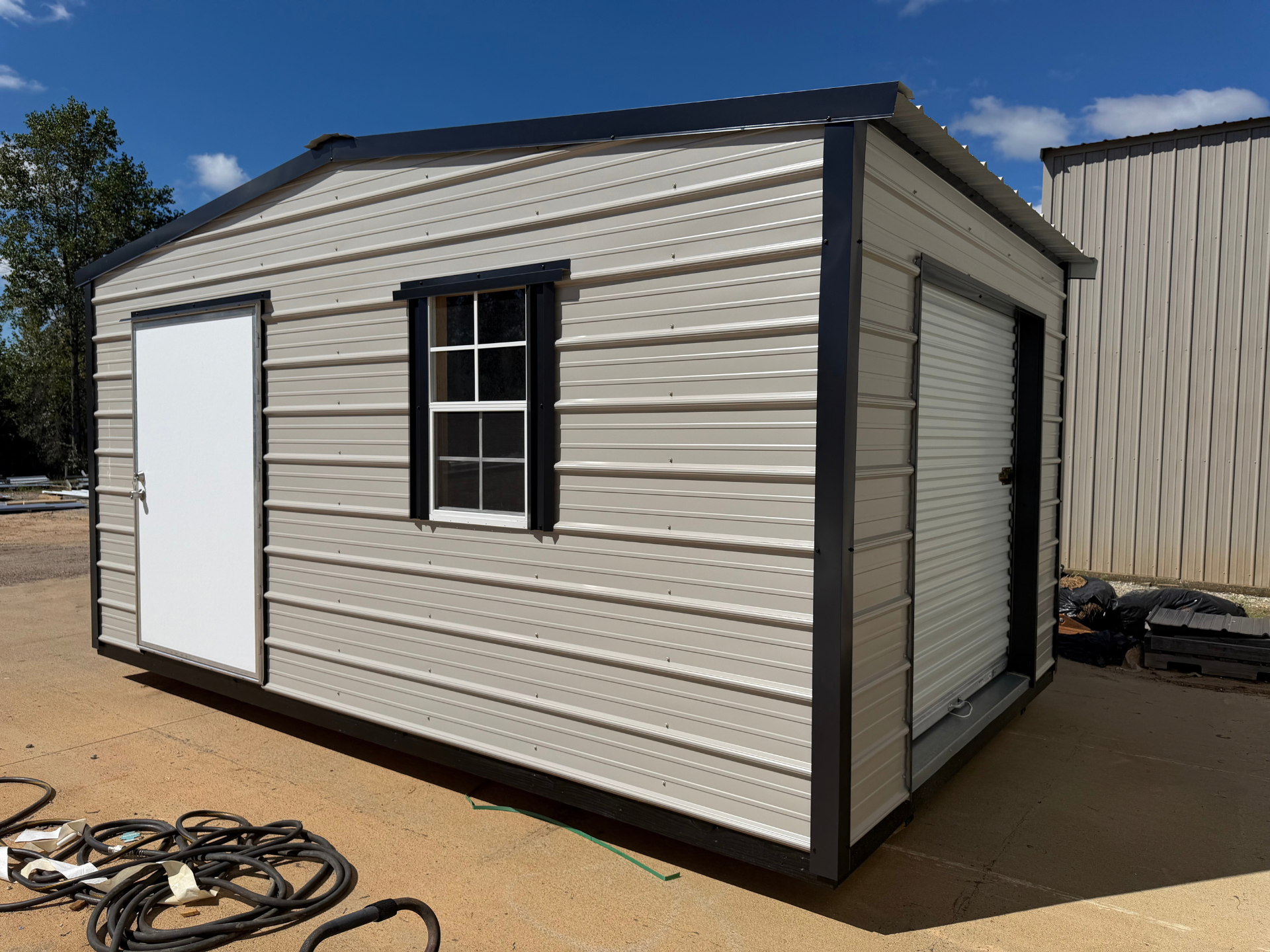 Small beige portable storage shed with white doors and black trim on a dirt lot outdoors