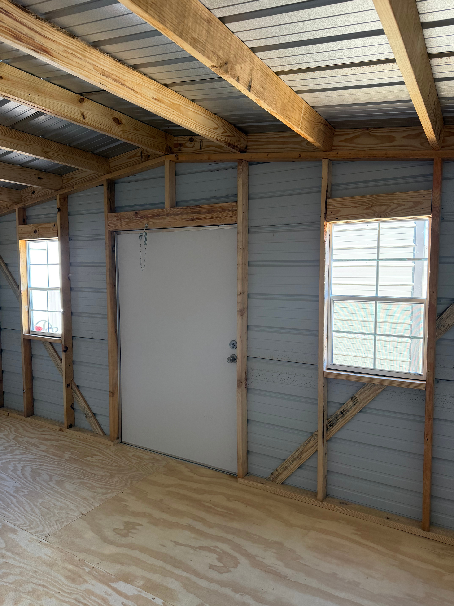 Interior of an unfinished room with exposed wood framing, plywood floor, and a white door between two windows.