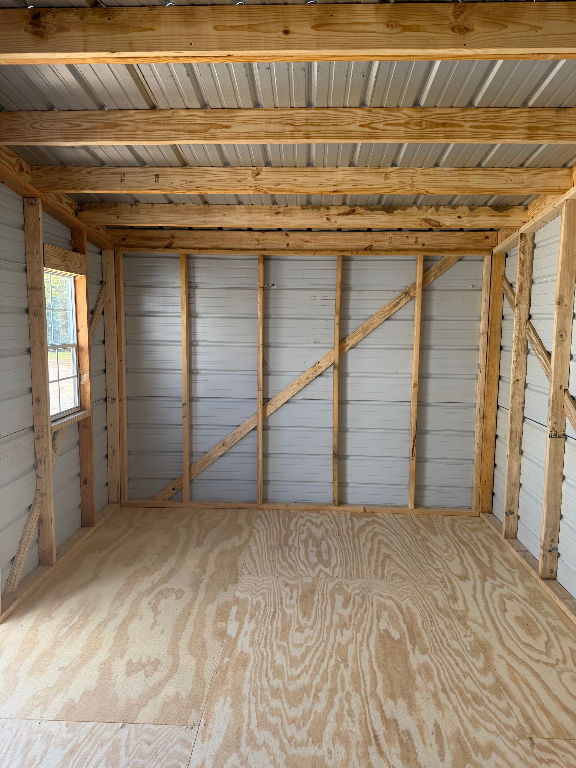 Empty wooden shed interior with unfinished plywood floor, exposed framing, and corrugated metal walls.