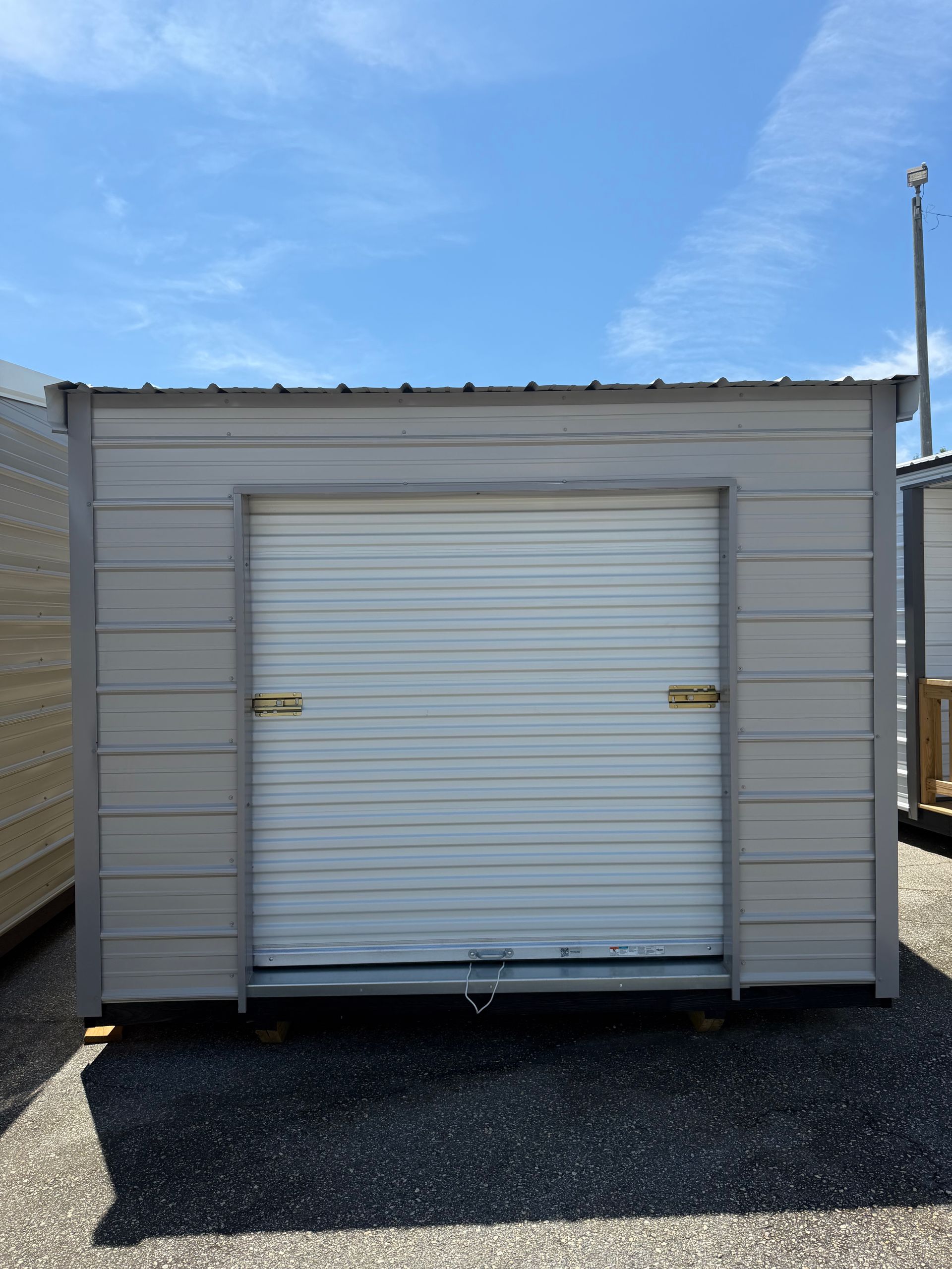 Small gray storage shed with a white roll-up door on a gravel lot under a blue sky