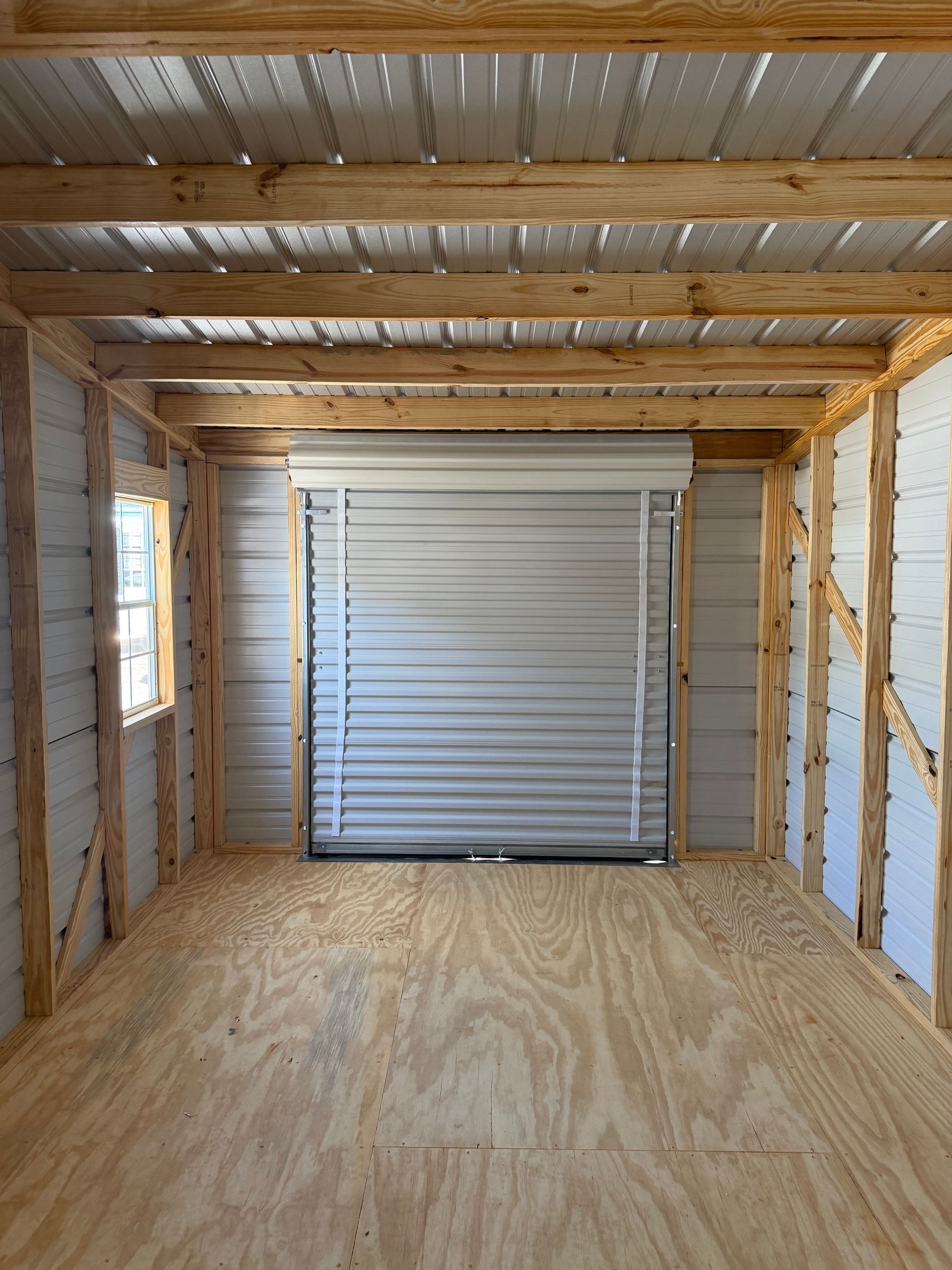 Empty unfinished garage interior with plywood floor, exposed studs, and a closed white roll-up door