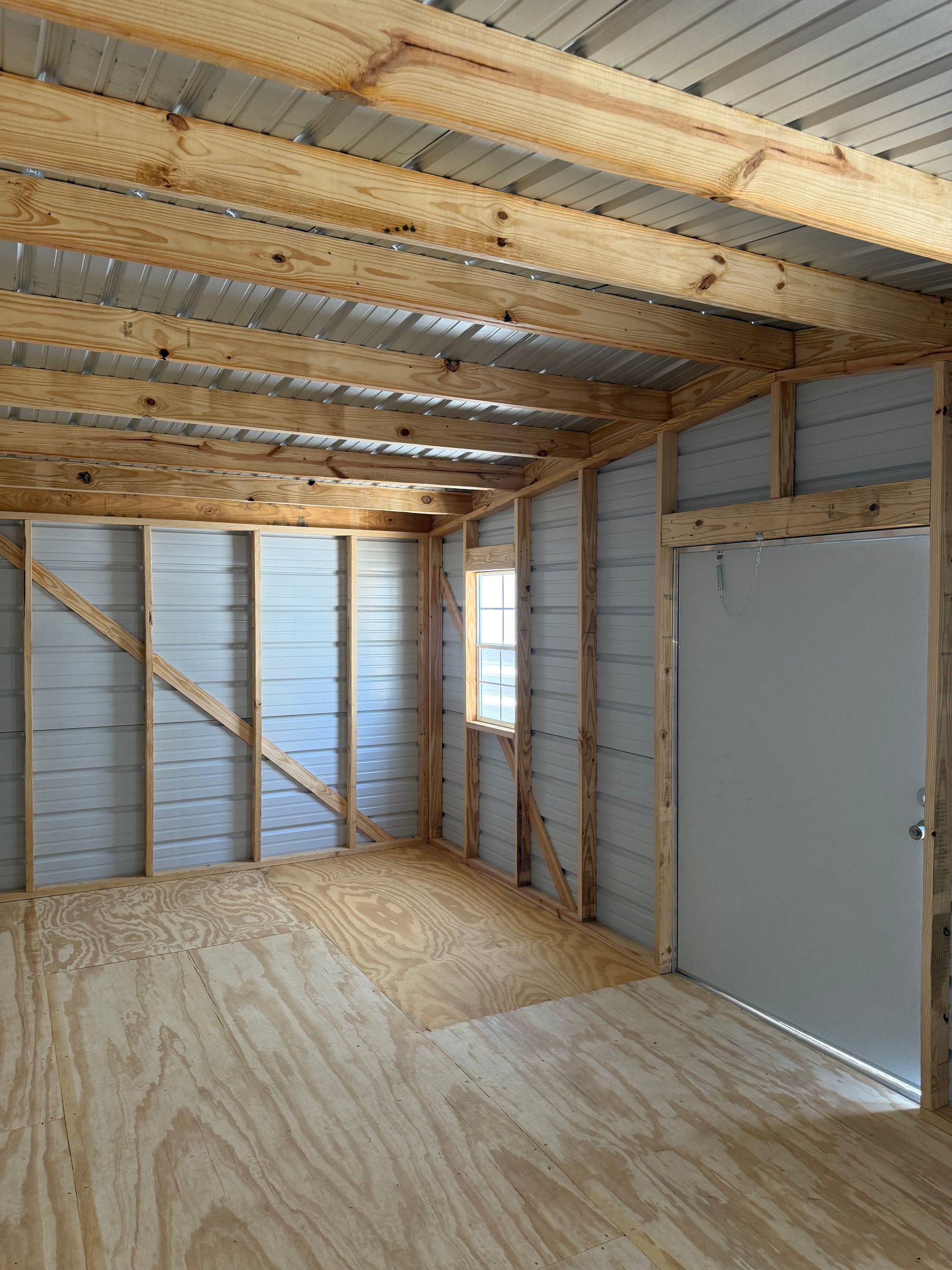 Unfinished room with plywood floor, exposed ceiling beams, insulation, and a white door on the right.