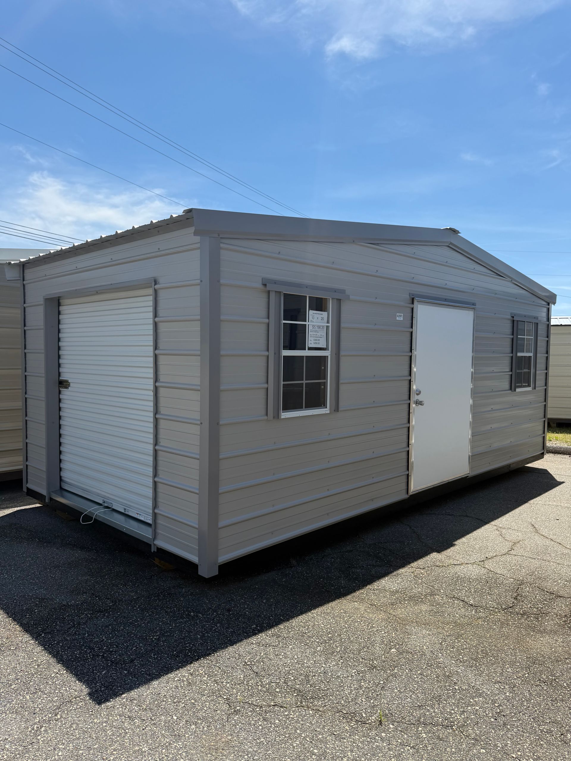 Gray portable shed with roll-up door and windows on a gravel lot under a blue sky
