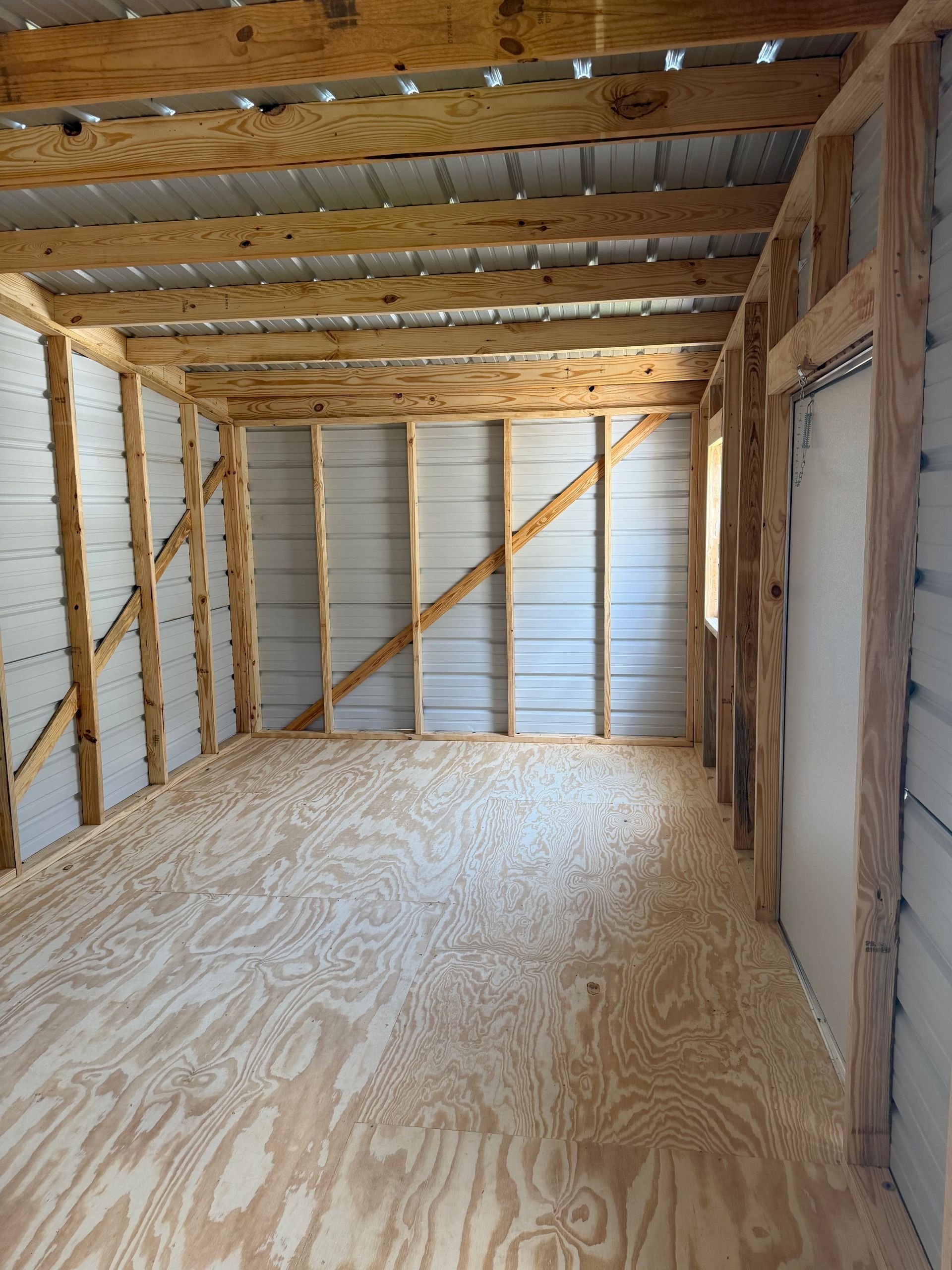 Empty unfinished room with exposed wood framing, plywood floor, and metal wall panels.