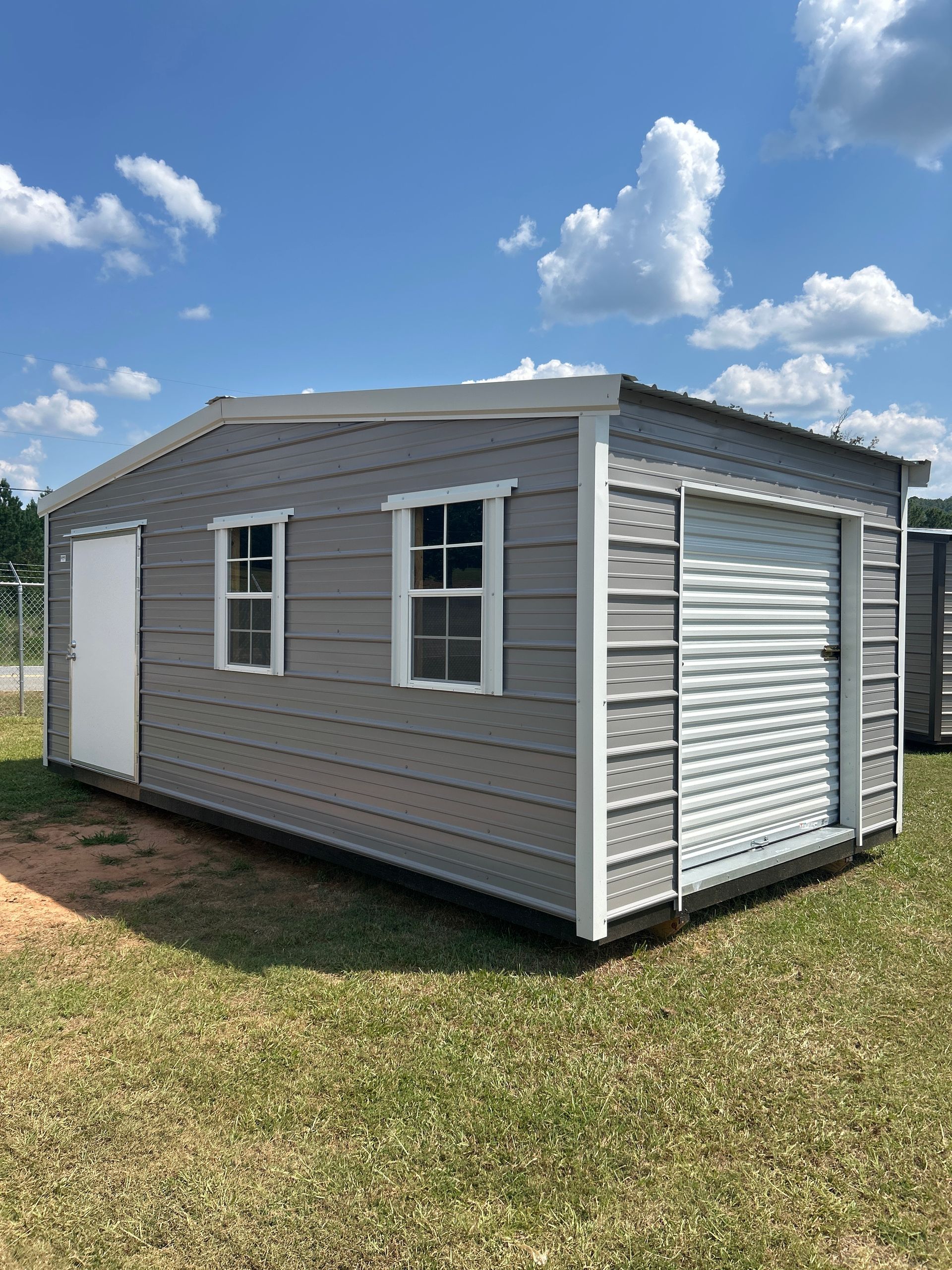 Gray shed with white door, windows, and garage door. Blue sky with clouds in background.