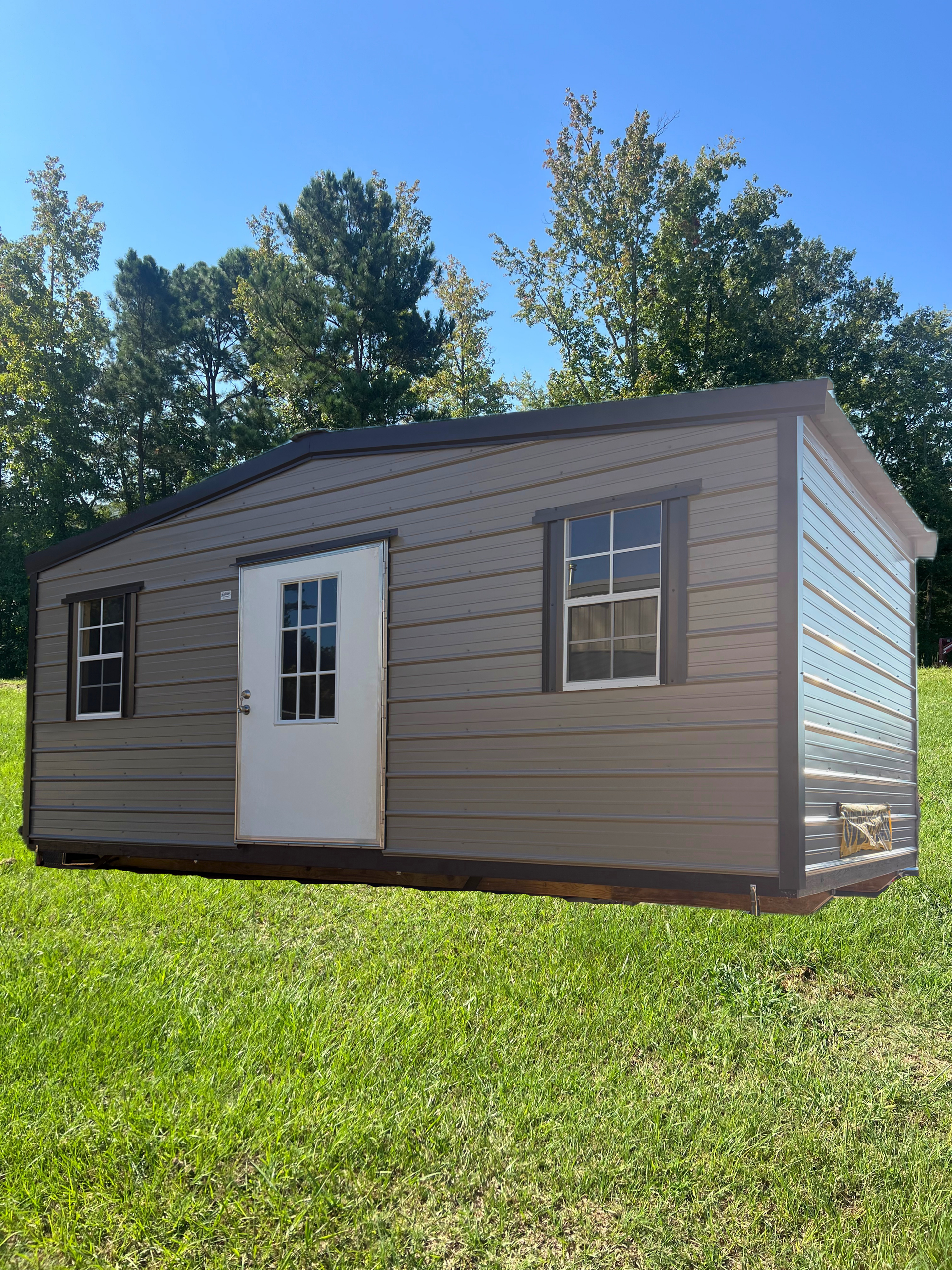 Small beige shed with white door and windows on a grassy lawn under a blue sky.