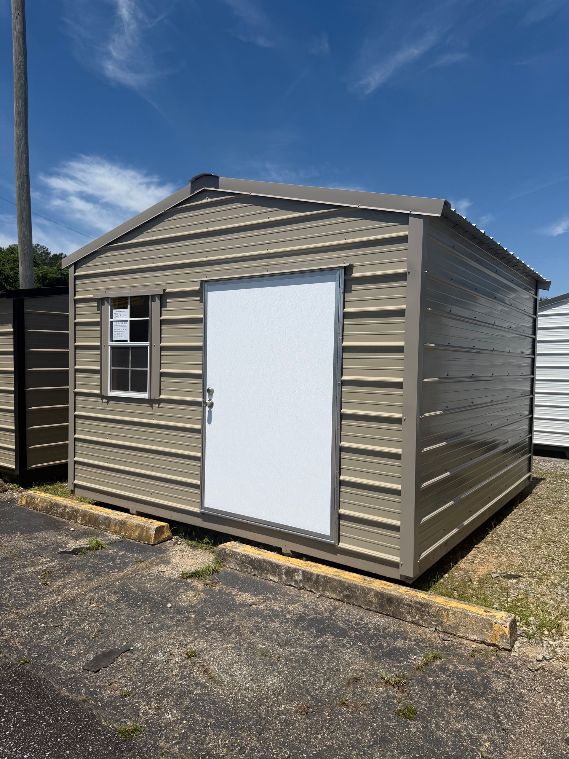 Small tan utility shed with a white door and window, sitting on a gravel lot under a blue sky.