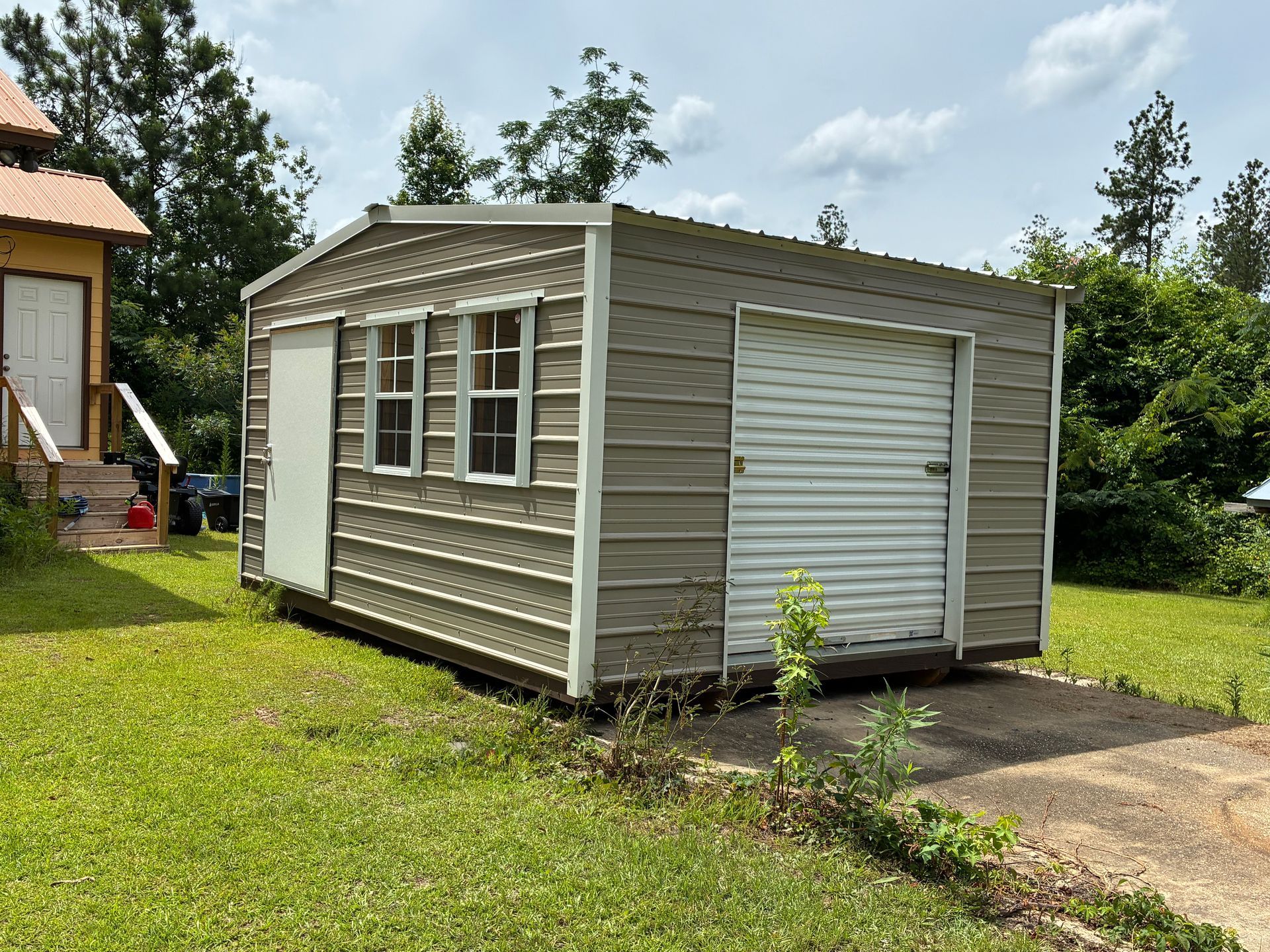 Small gray shed with white-trimmed windows and a roll-up door beside a grassy yard and driveway
