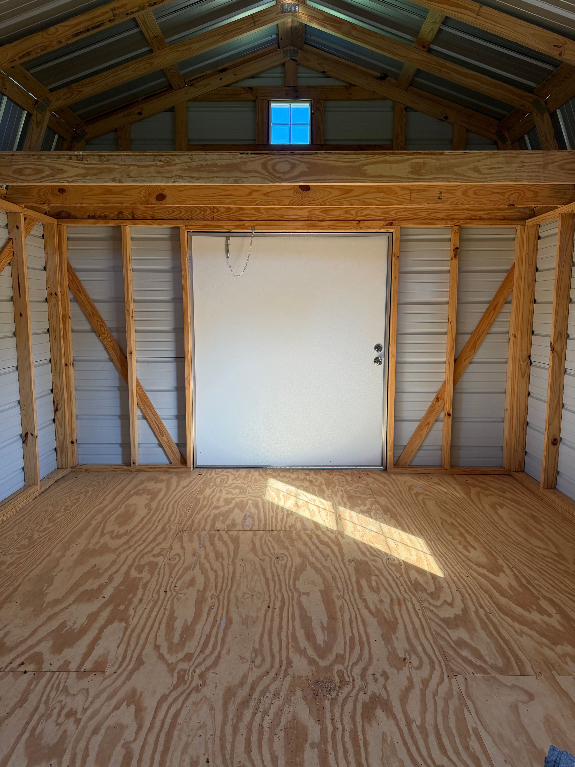 Empty wooden attic with exposed beams, plywood floor, and a white door under a blue skylight.