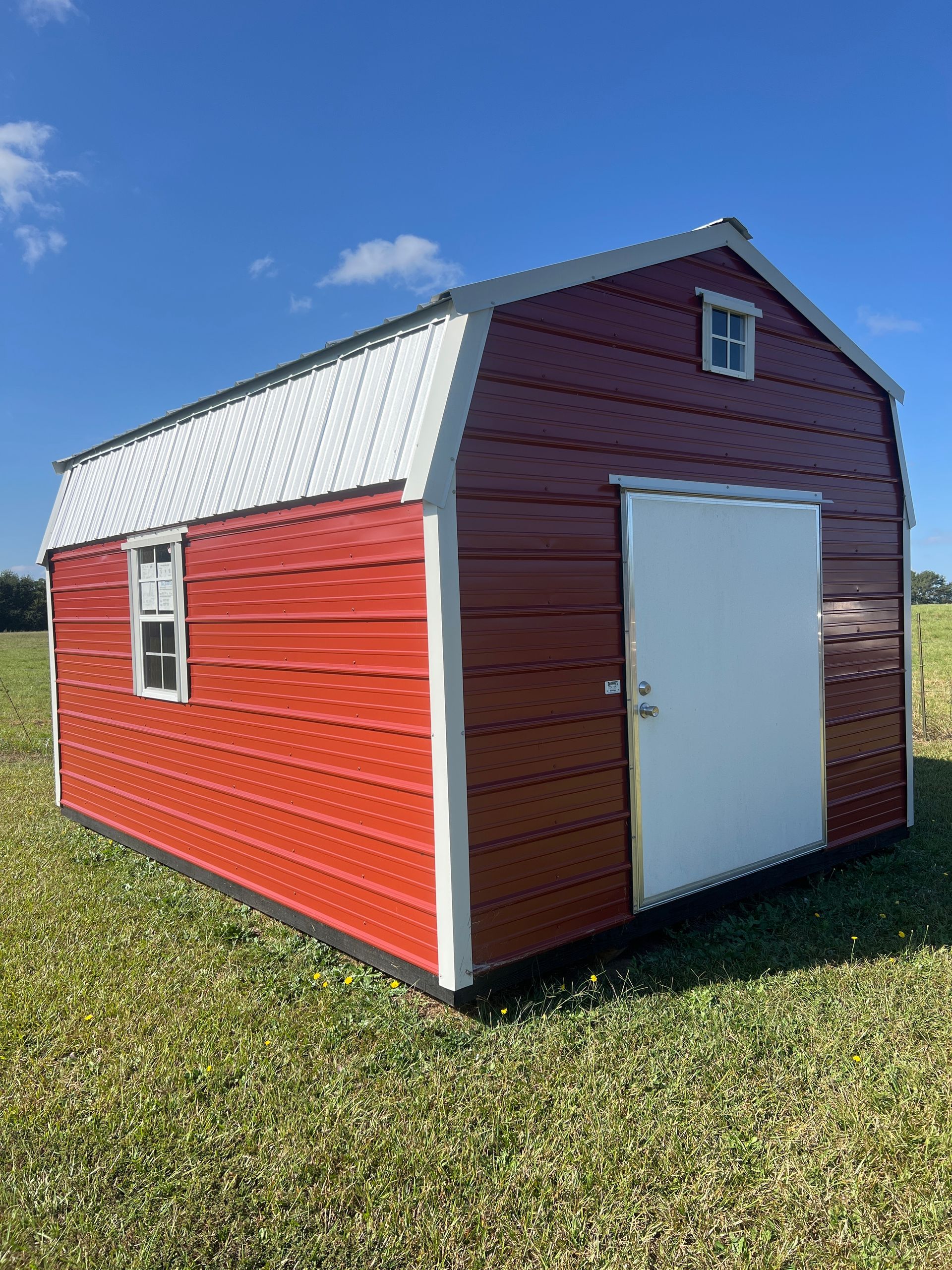 Red and white shed with a metal roof sitting in a grassy field under a blue sky.