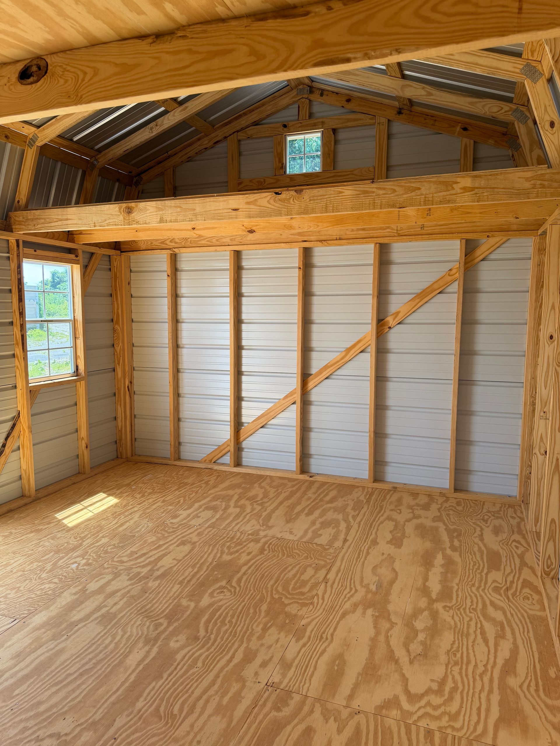 Unfinished wooden shed interior with plywood floor, exposed rafters, and framed walls with windows and insulation view.