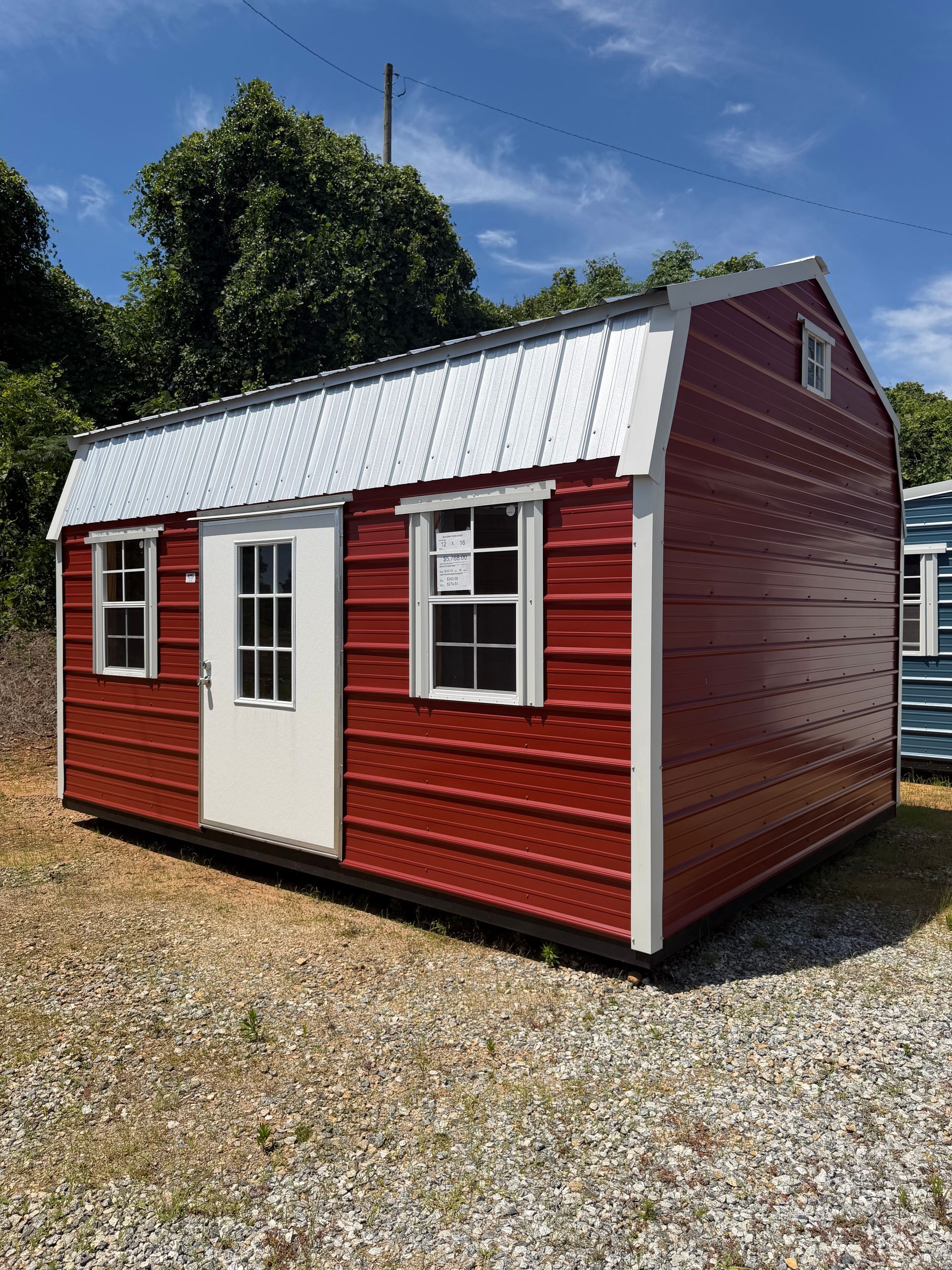 Small red and white shed with a metal roof on a gravel lot under a blue sky