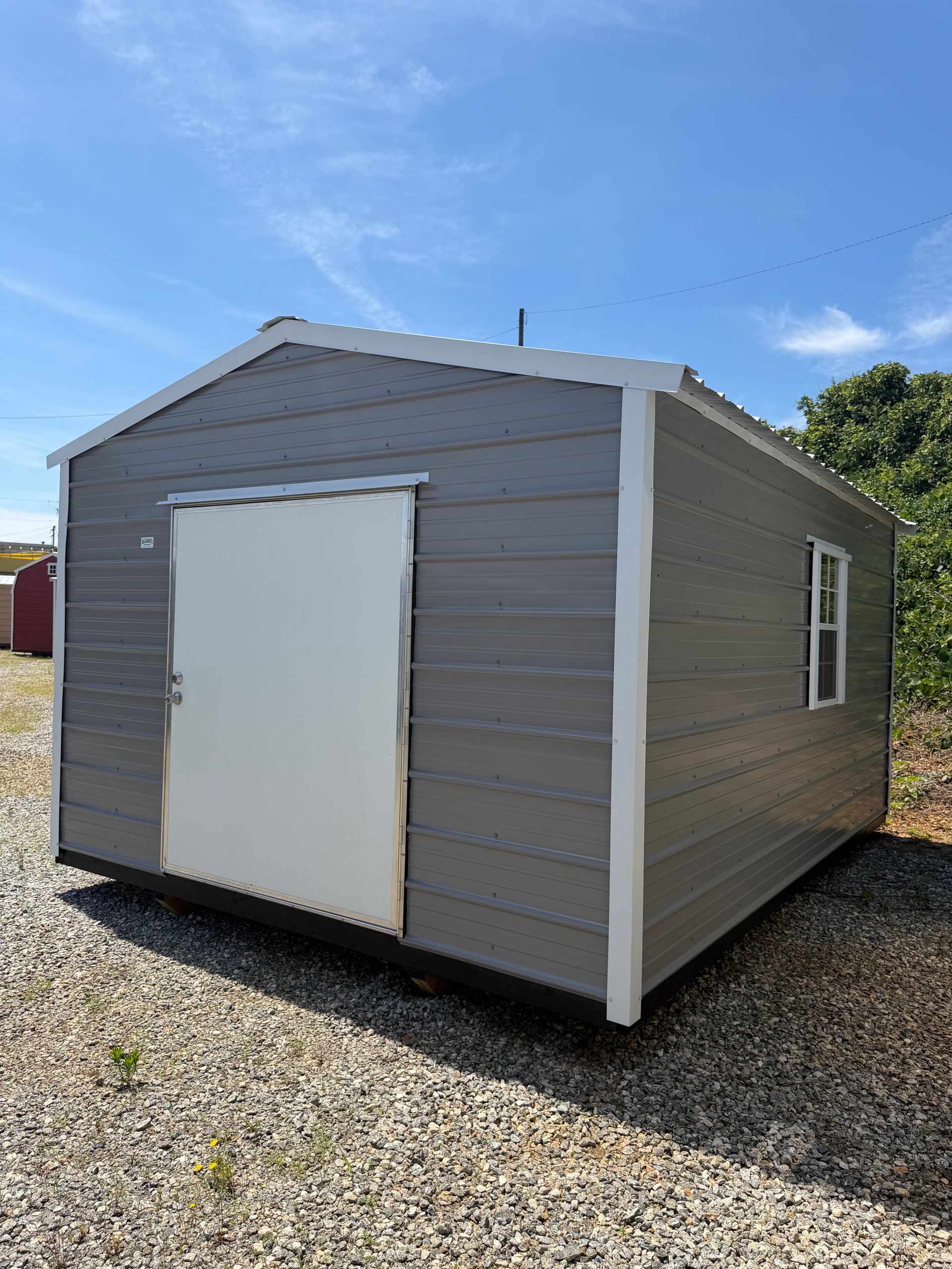 Small gray shed with white trim and a white door, sitting on a gravel lot under a blue sky.