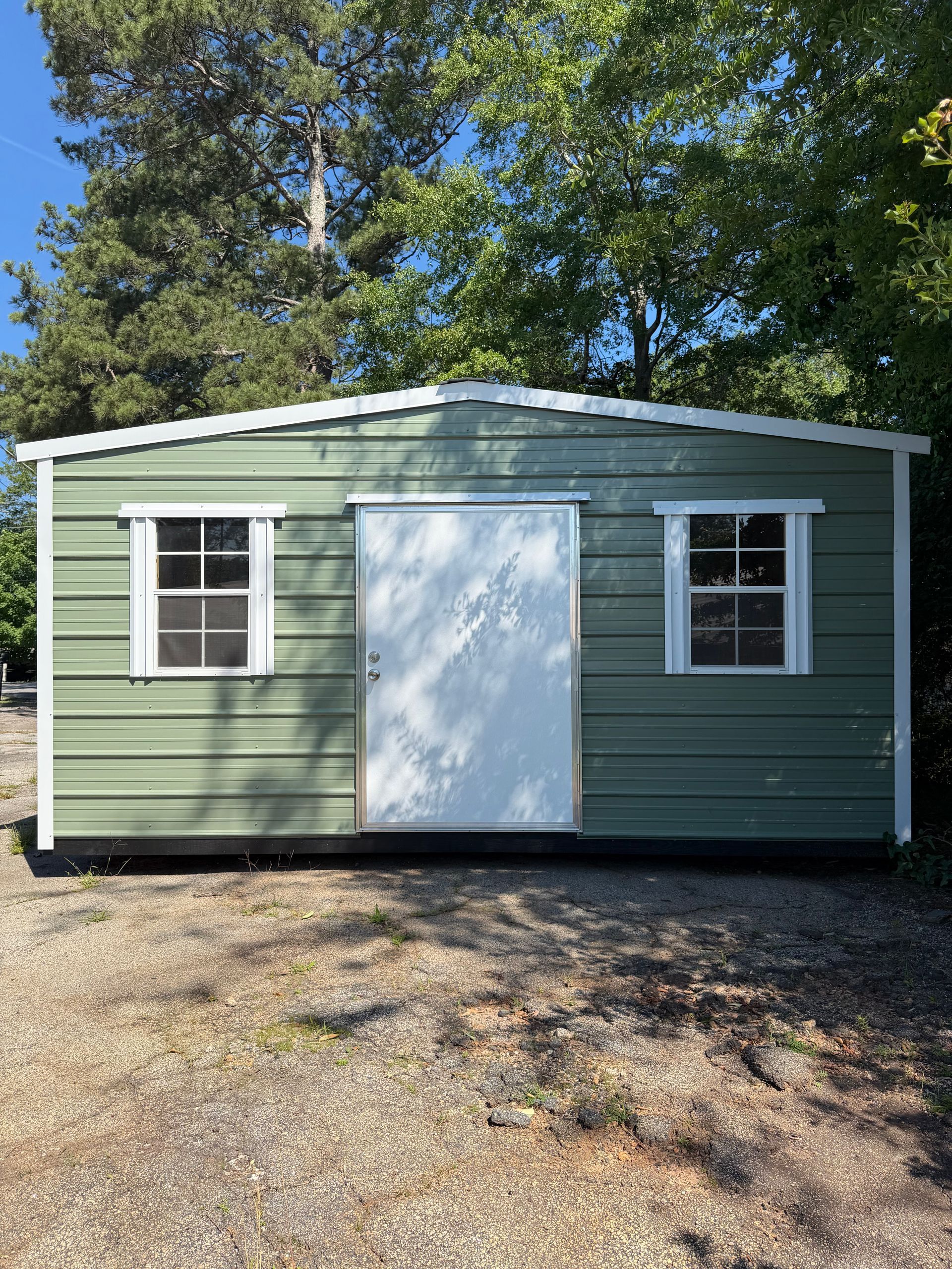 Small green shed with white door and windows, set on gravel beneath tall trees.