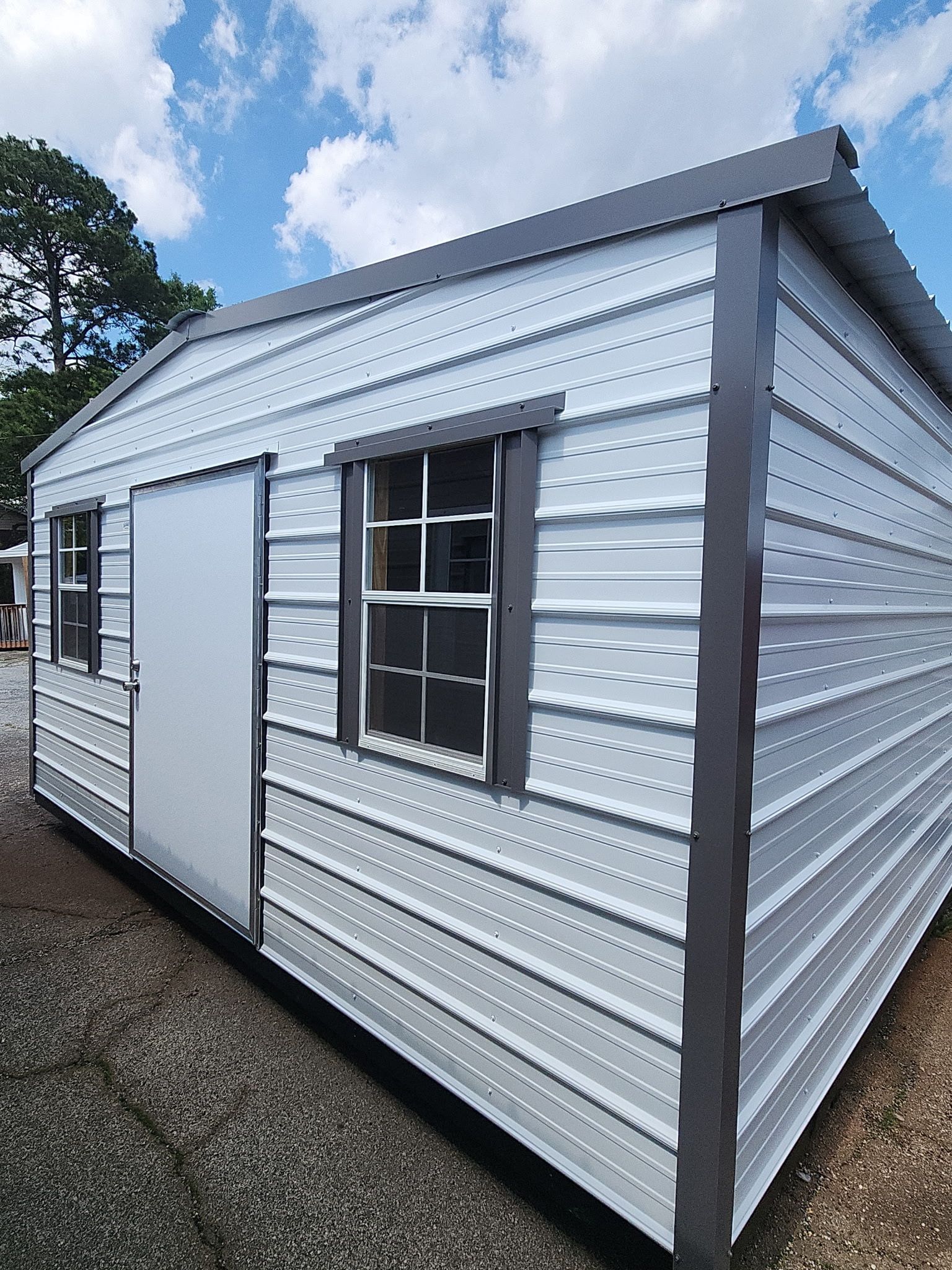 Silver metal shed with a window and side door on a gravel lot under a cloudy sky