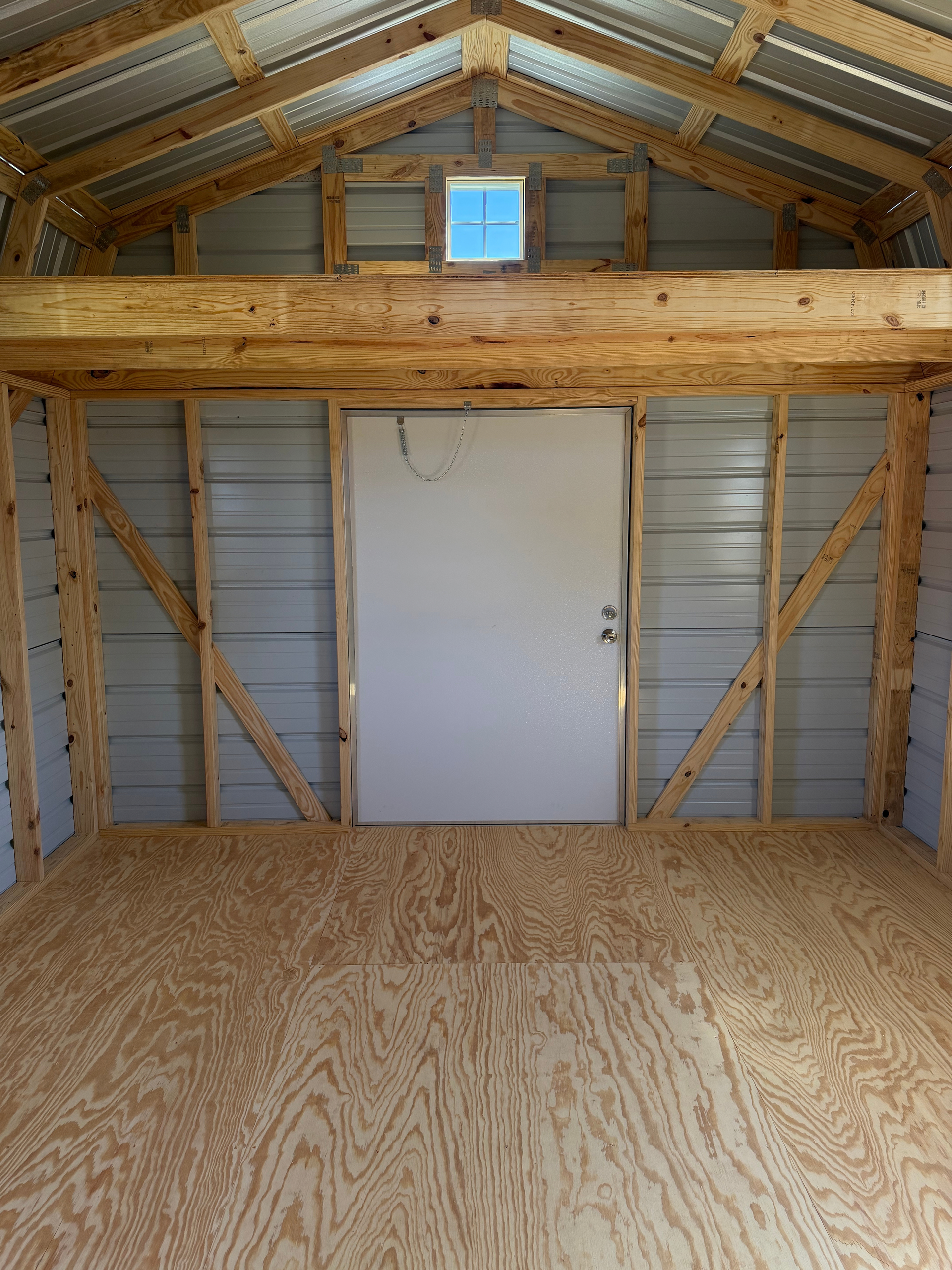 Empty unfinished shed interior with plywood floor, metal walls, and a white door centered on the back wall