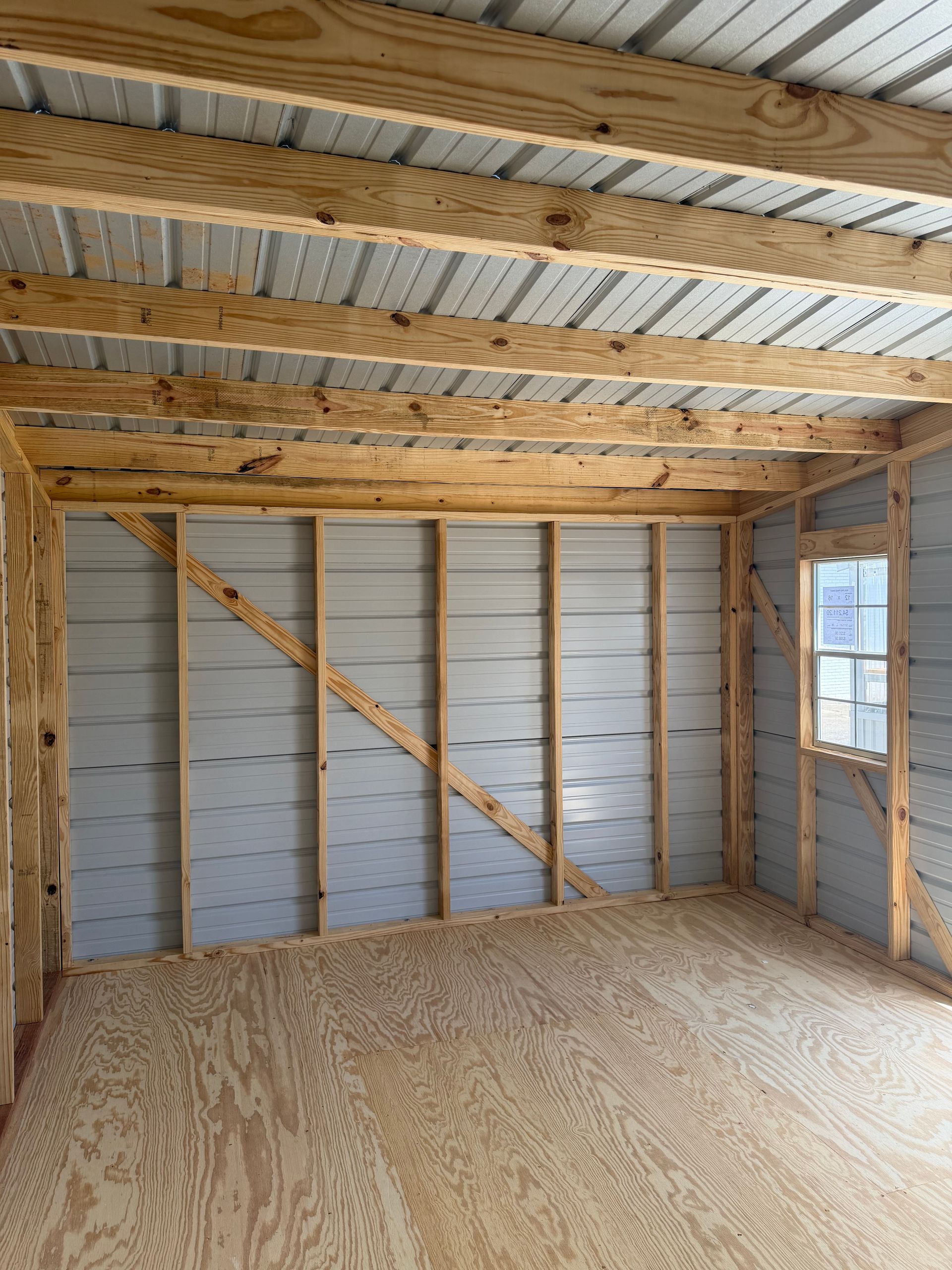 Unfinished wooden room interior with exposed ceiling beams, plywood floor, and a small window.