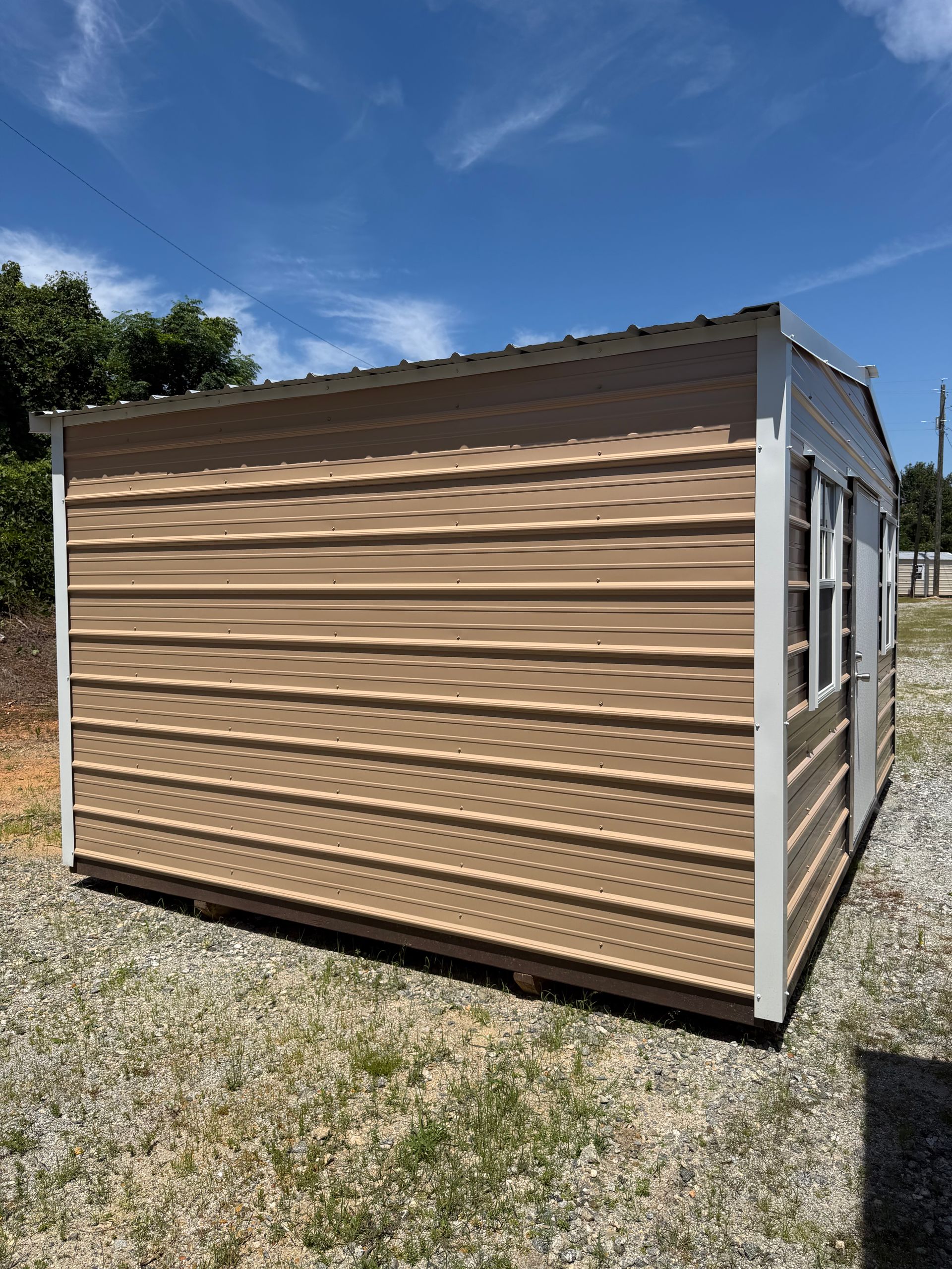 Tan mobile shed with tan siding and white trim on a gravel lot under a blue sky