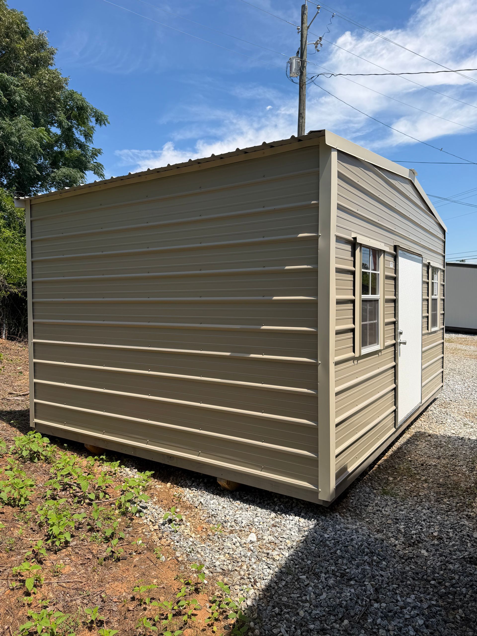 Tan shed with white trim and windows beside a gravel driveway on a sunny day