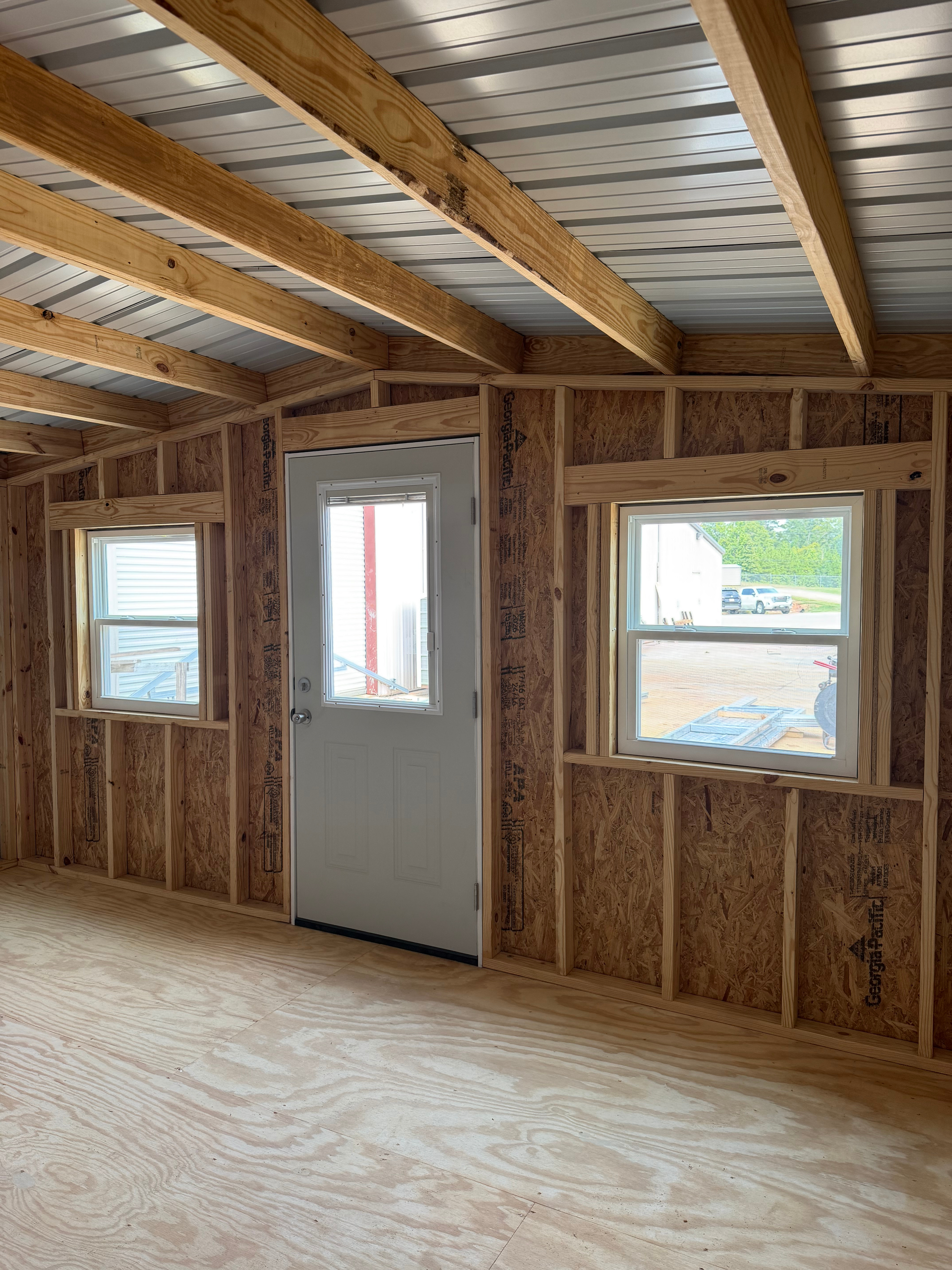 Unfinished room with exposed wood framing, plywood floor, and three windows on one wall.