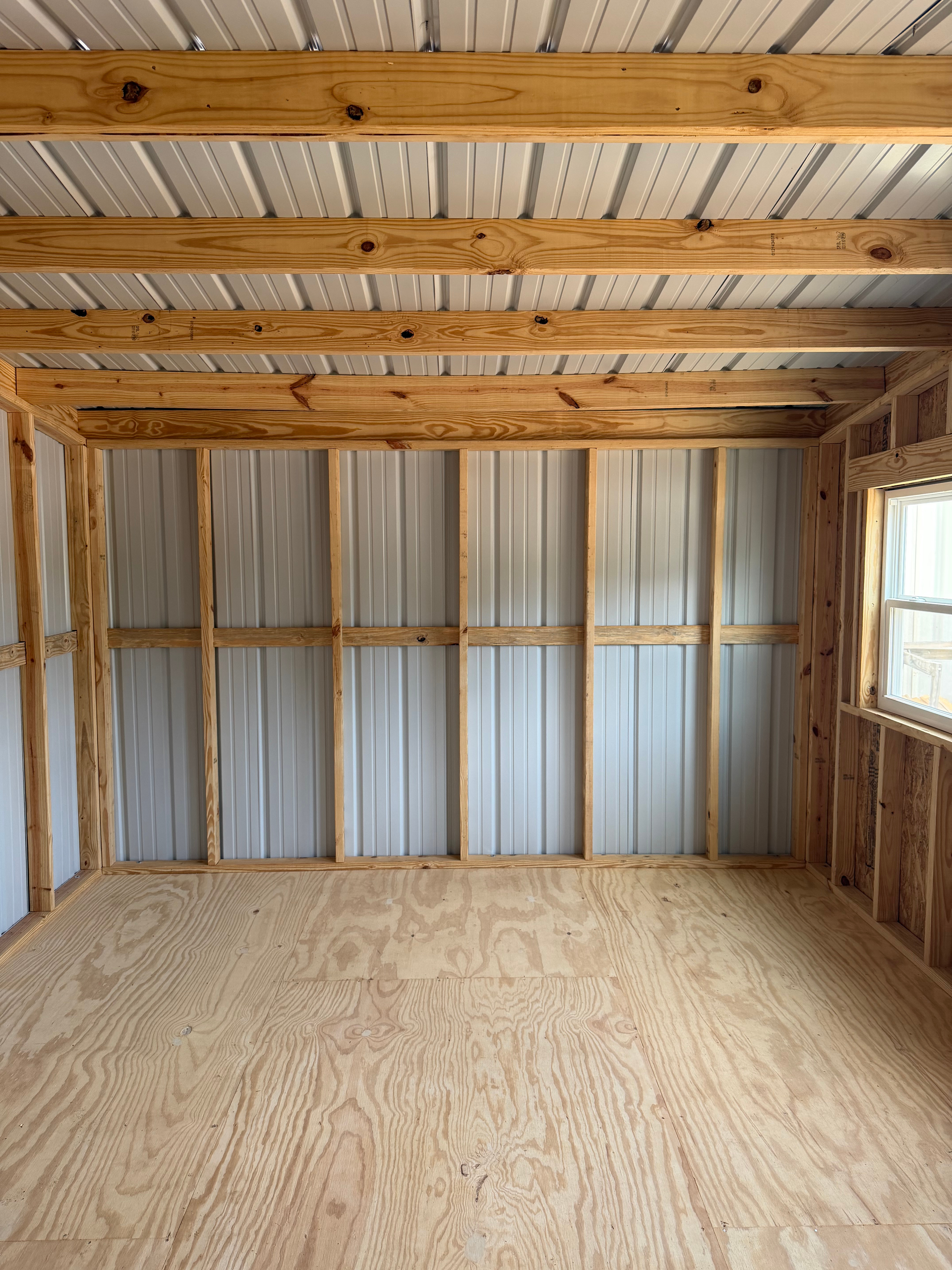 Empty unfinished shed interior with plywood floor, exposed wood beams, and corrugated metal wall panels.