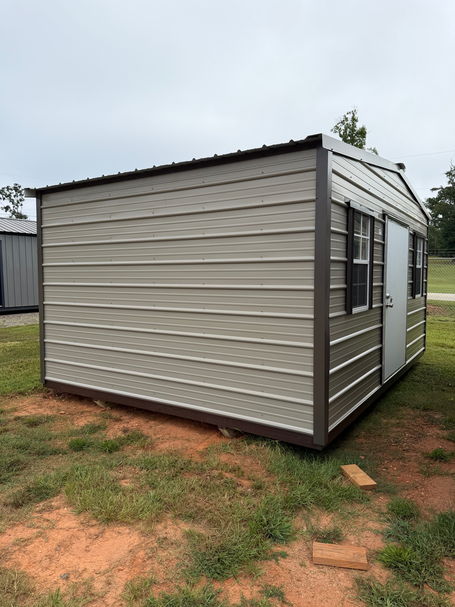Small beige portable cabin with windows on a grassy dirt lot, viewed from the corner.