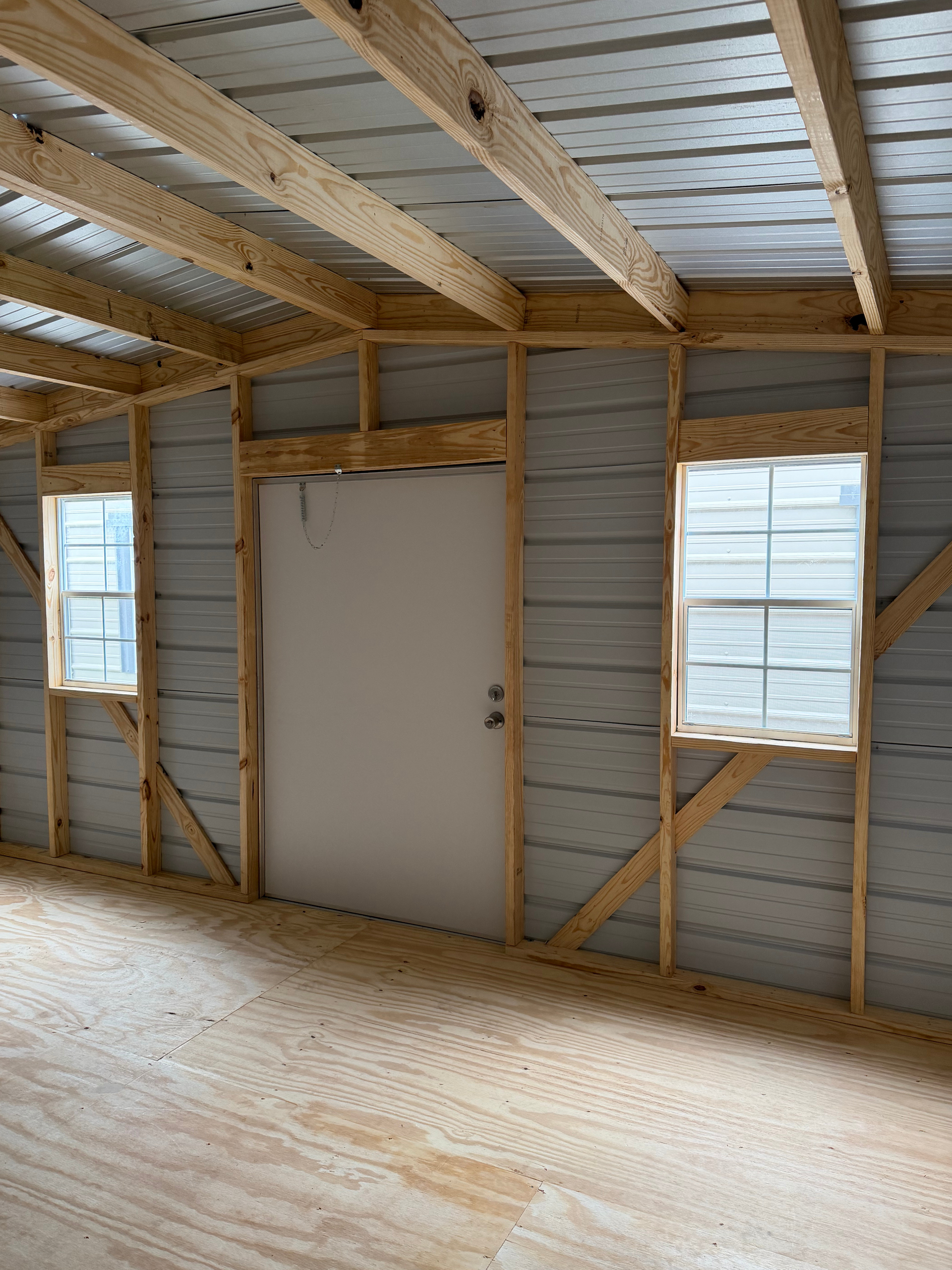 Unfinished room with exposed wood framing, corrugated metal walls, two windows, and a white door.