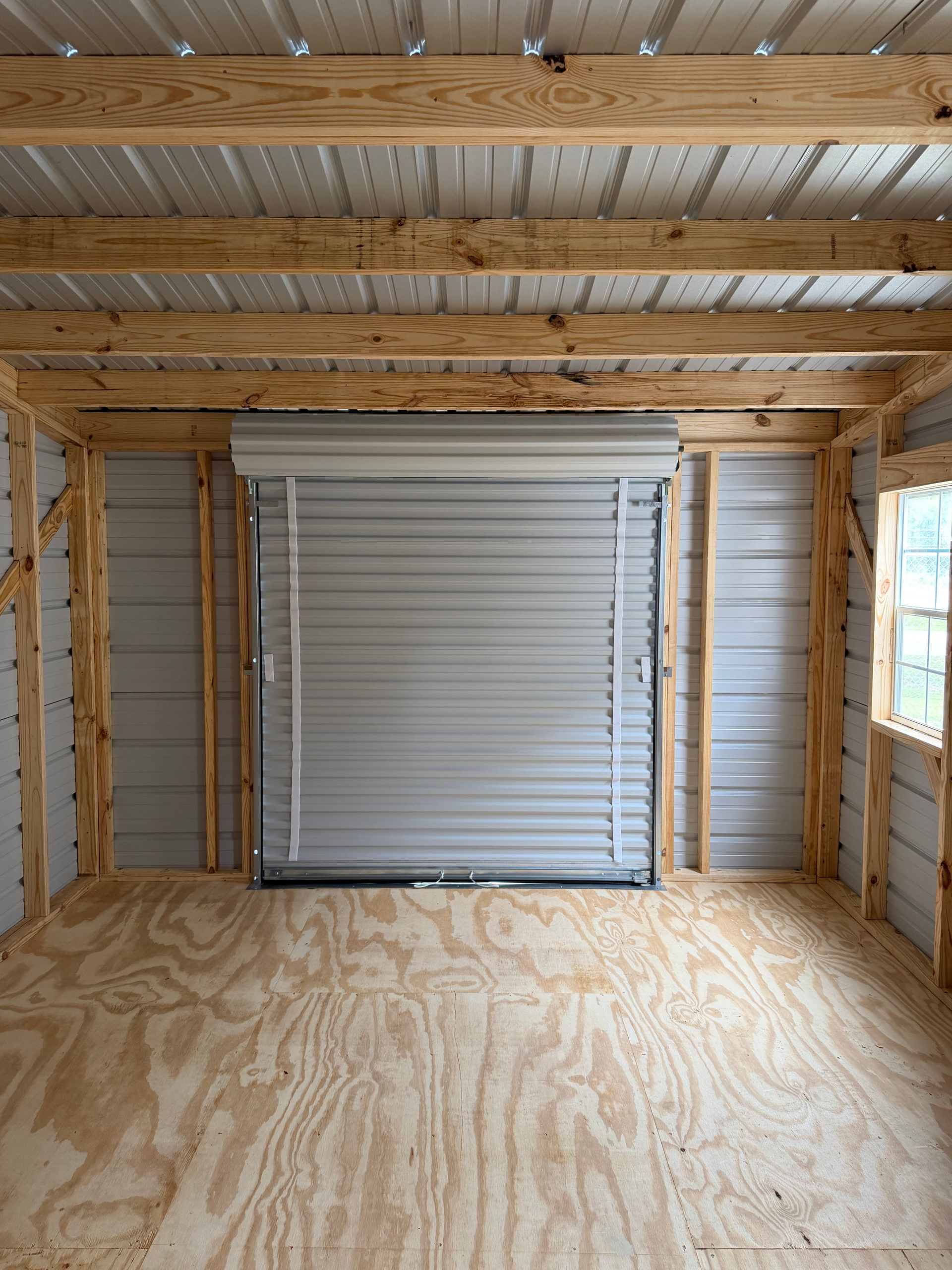 Empty wooden garage with exposed beams, plywood floor, and a closed white roll-up door.