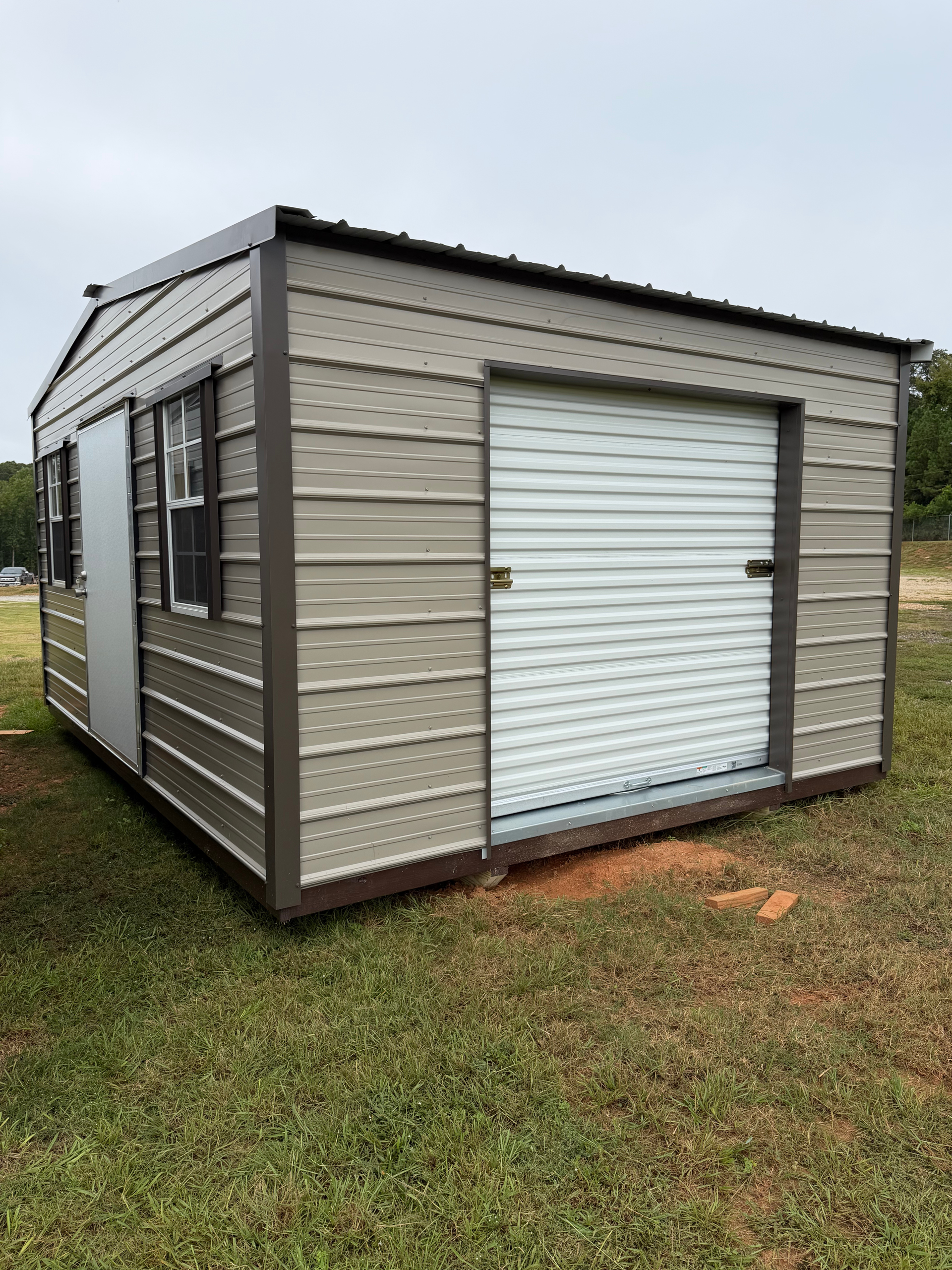 Small beige storage shed with a white roll-up door on grass under an overcast sky