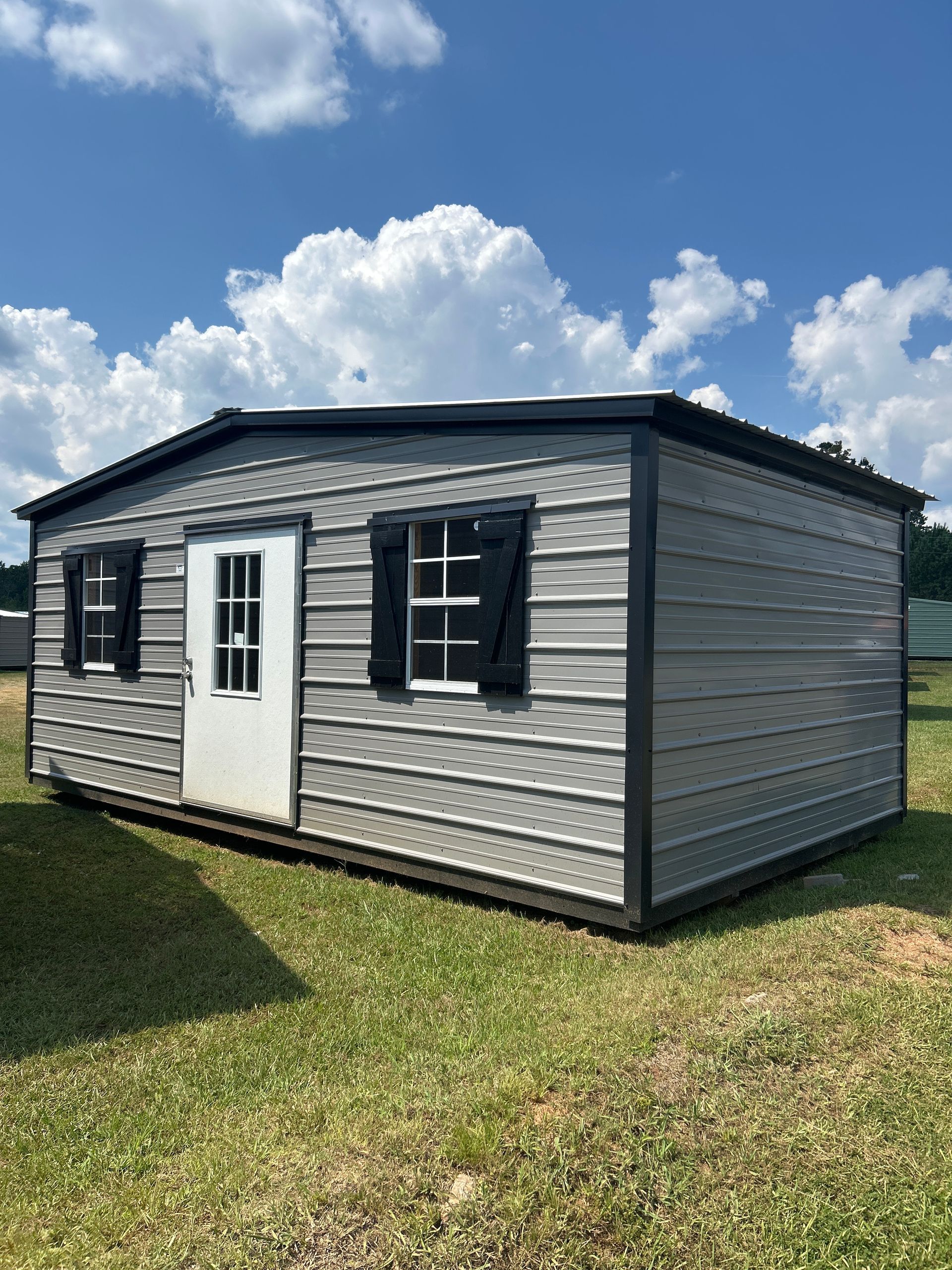 Gray metal shed with a white door, black shutters, and trim, set in a grassy area with blue sky.