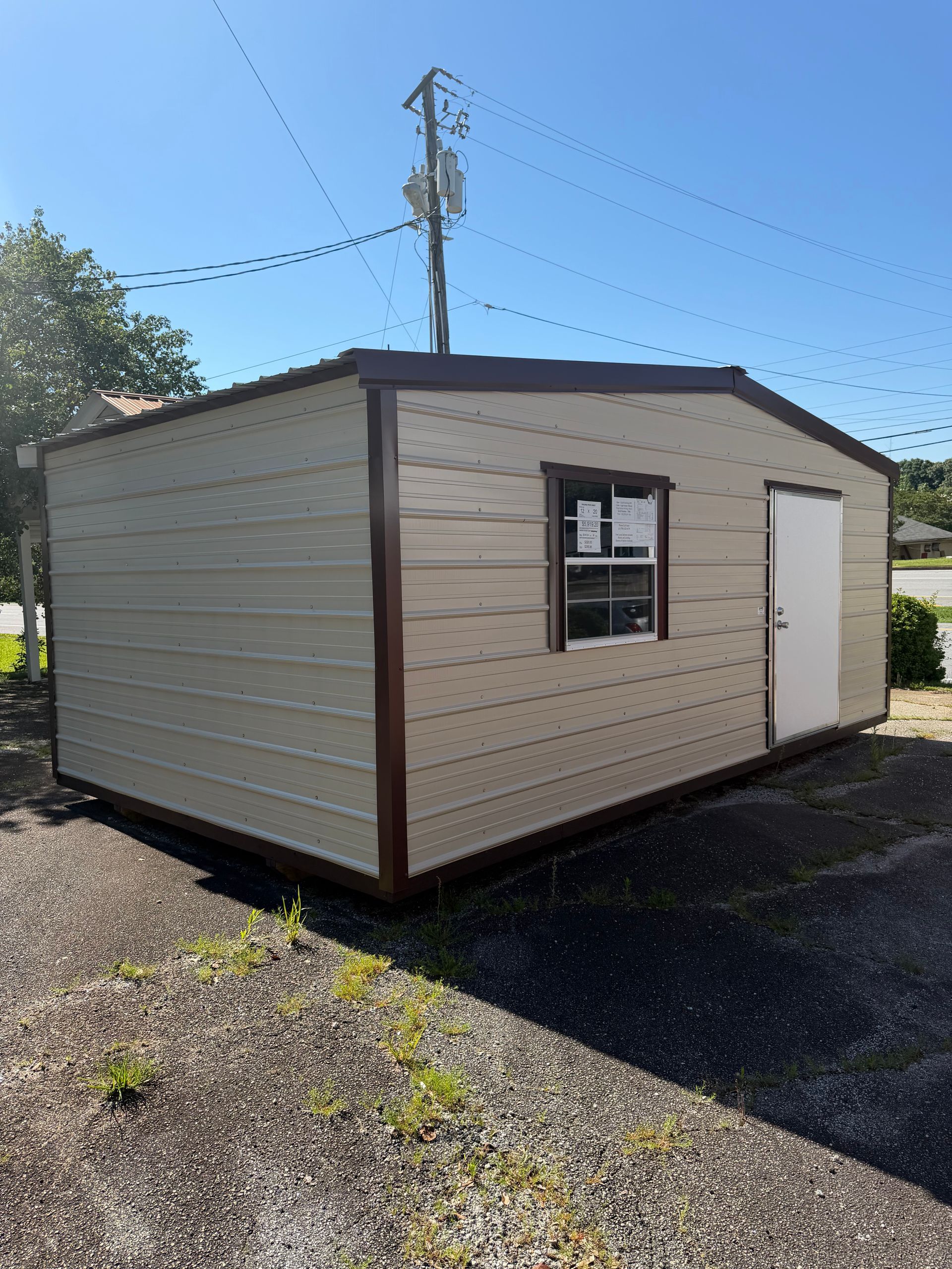 Small beige shed with brown trim, a window, and white door on a gravel lot under a clear blue sky