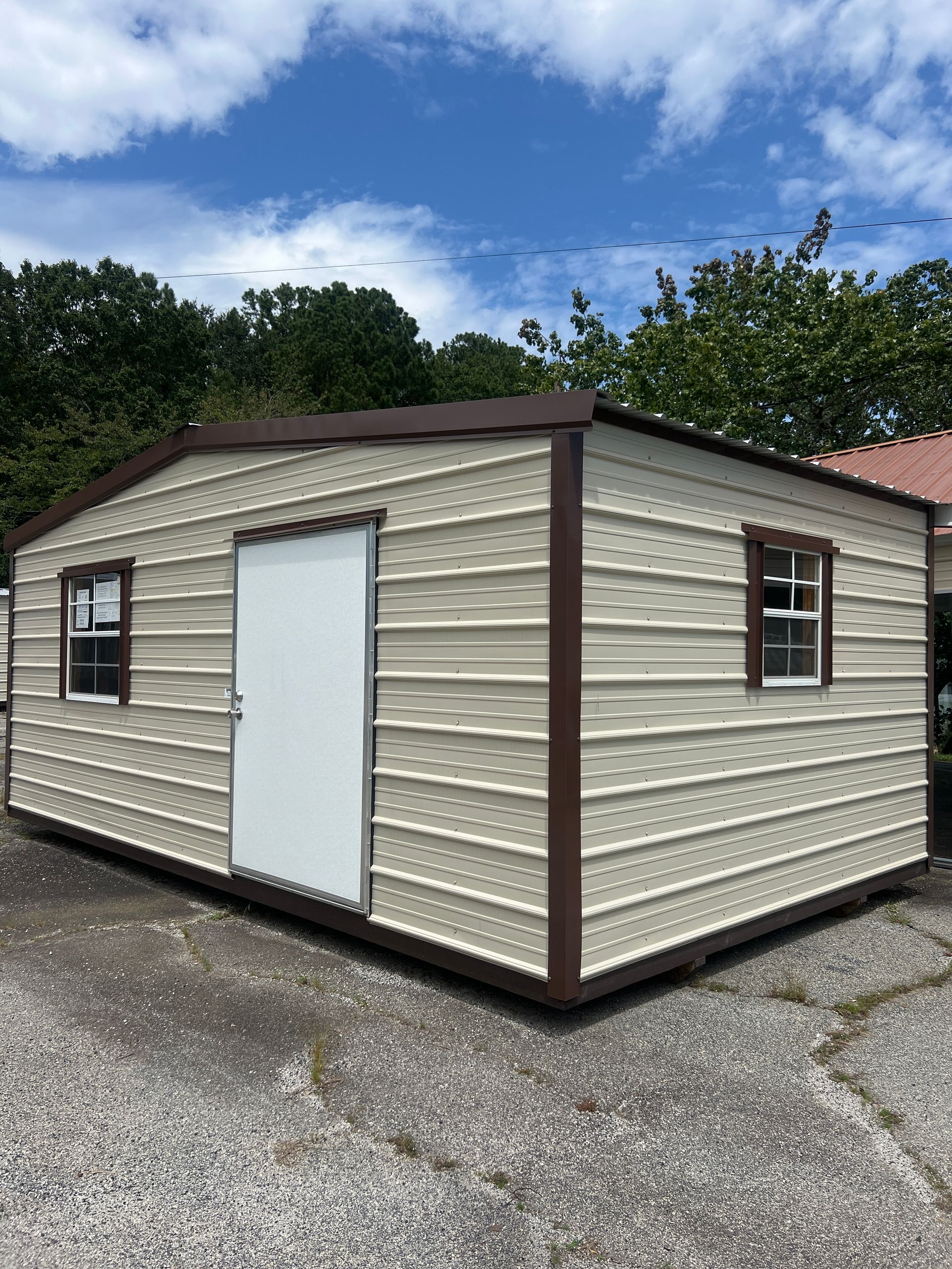 Small beige shed with white door and brown trim on cracked pavement under a cloudy sky