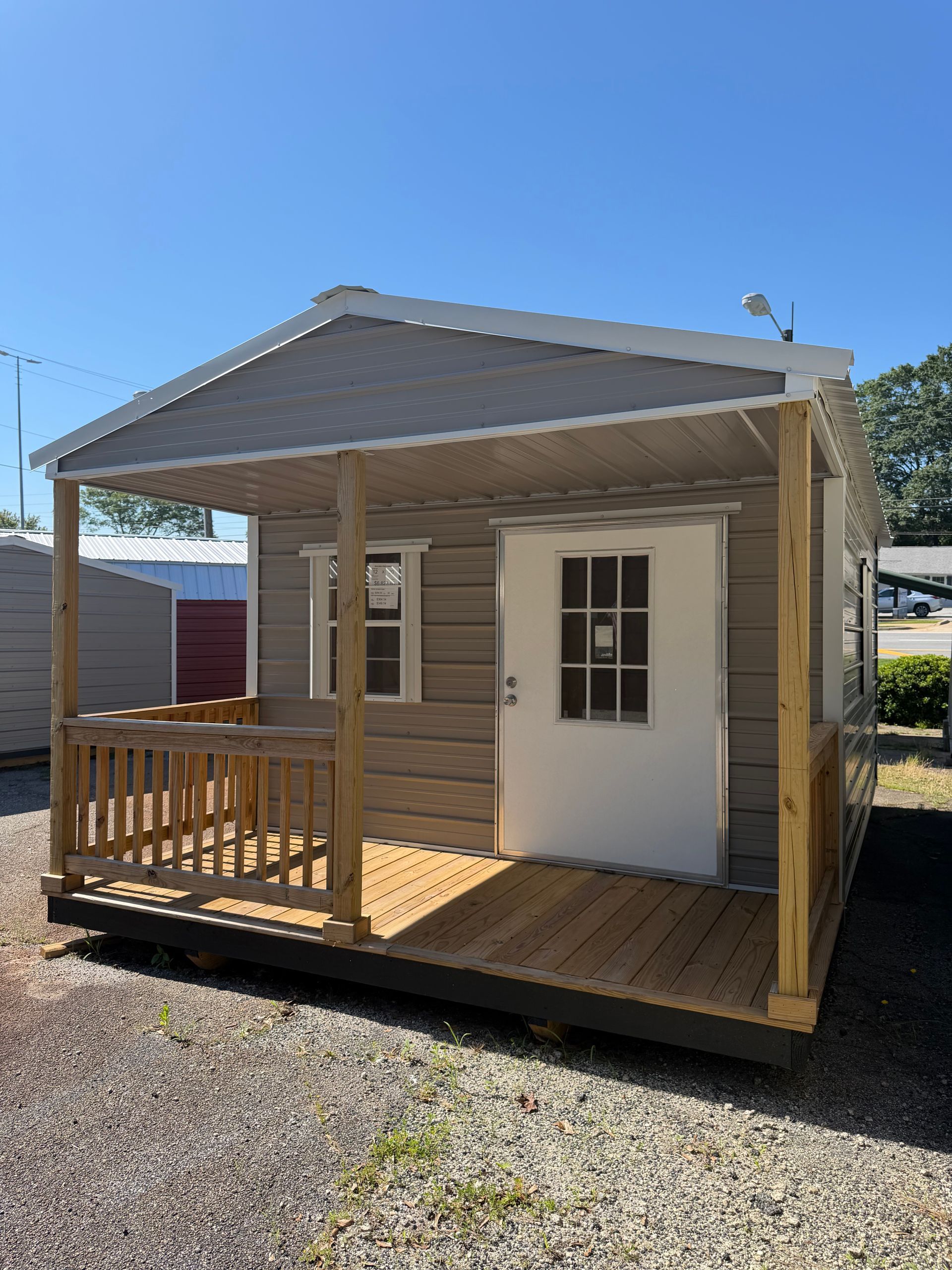 Small beige wooden cabin with porch and white door on a gravel lot under a clear blue sky
