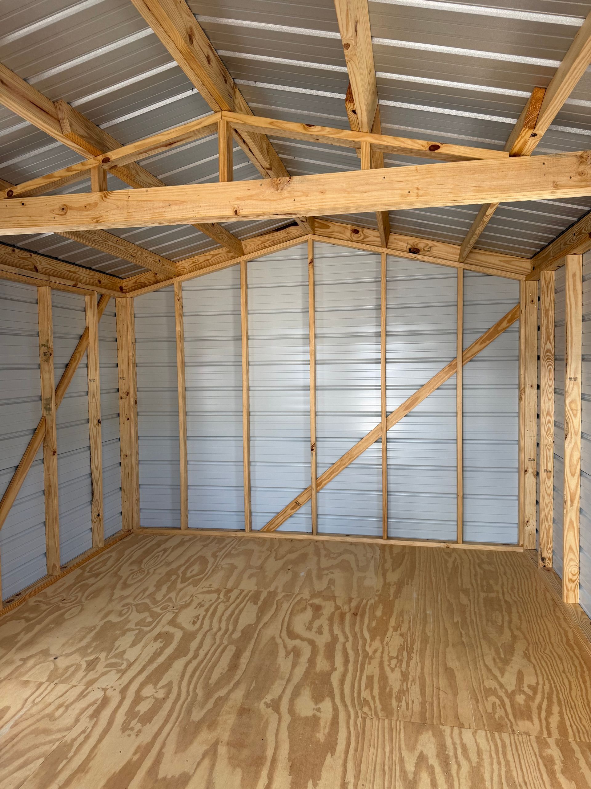 Empty unfinished shed interior with plywood floor, exposed wood framing, and metal roof panels.