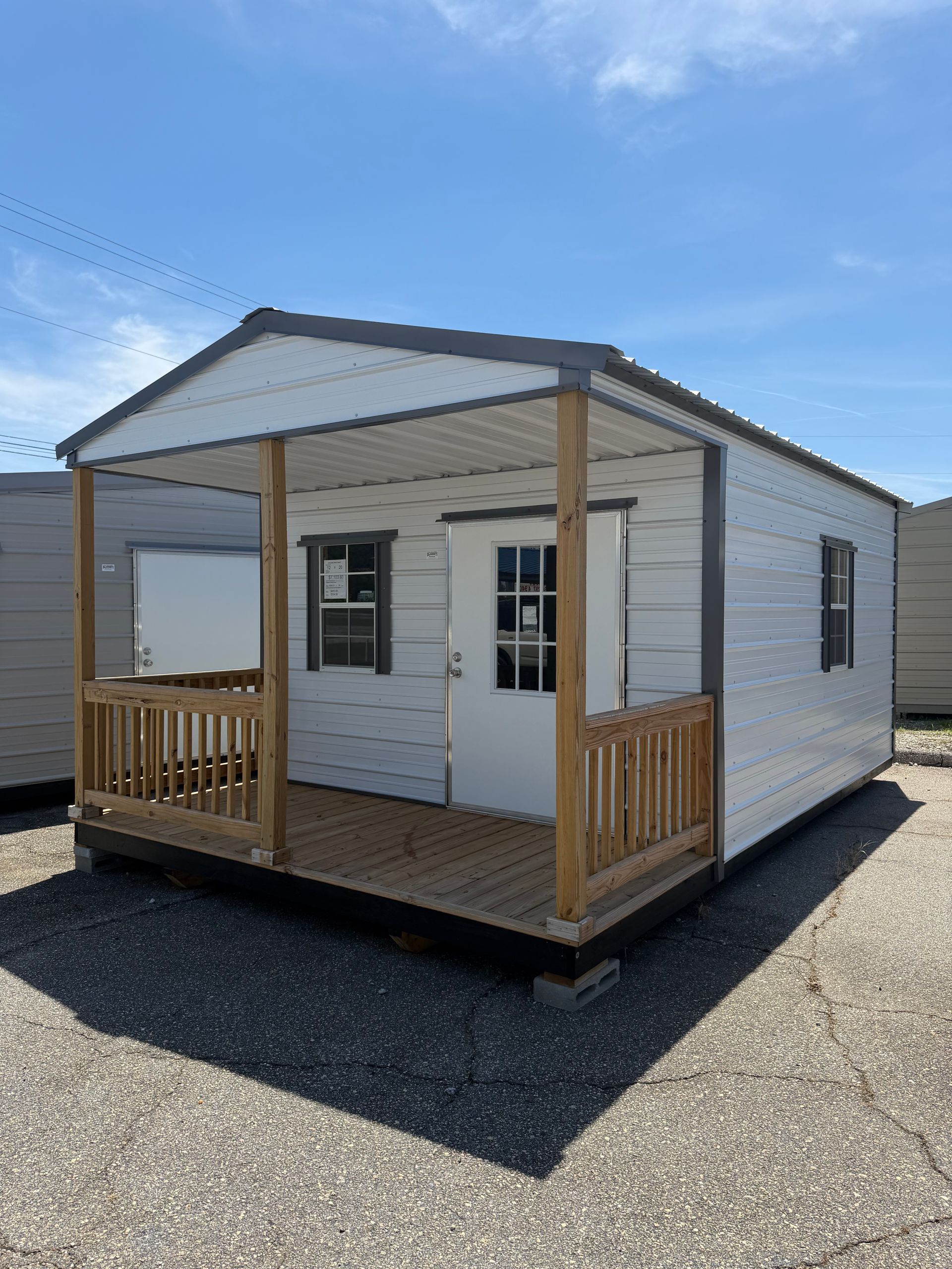Small white tiny house with a front porch and gray roof on a gravel lot under a blue sky