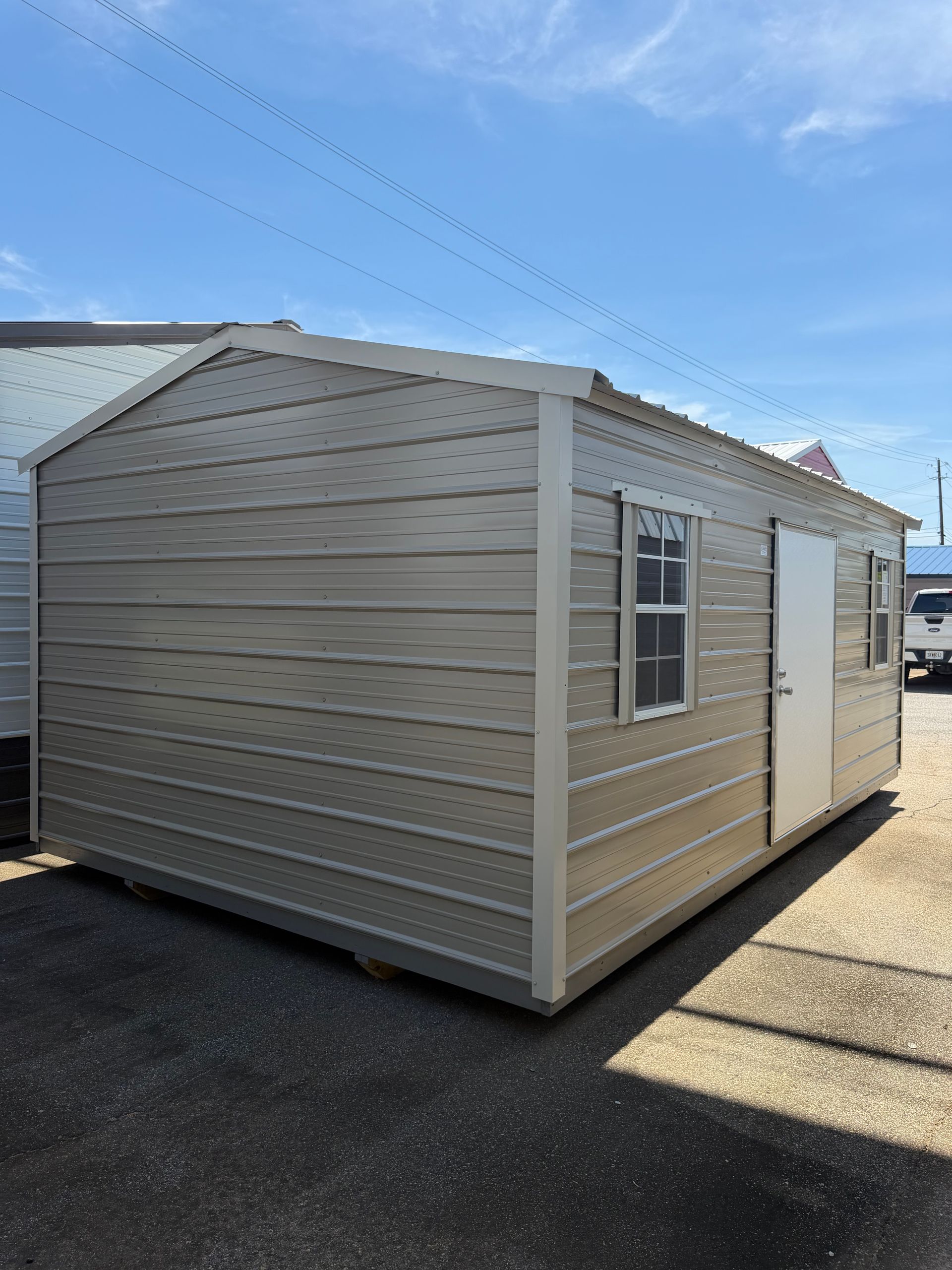 Beige portable shed with white trim and windows on an asphalt lot under a blue sky