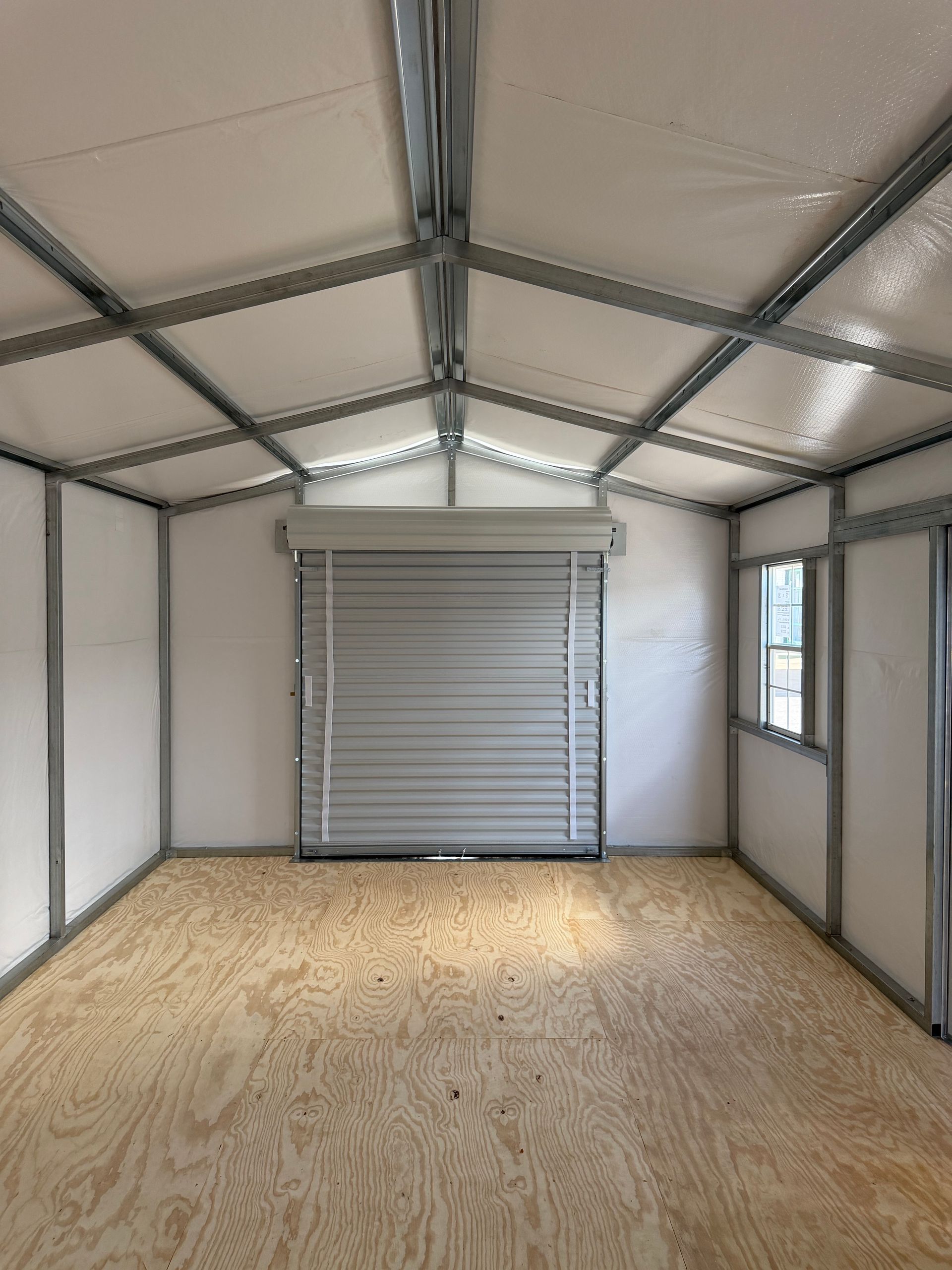 Interior of an unfinished wooden room with exposed framing, plywood floor, and a small window.