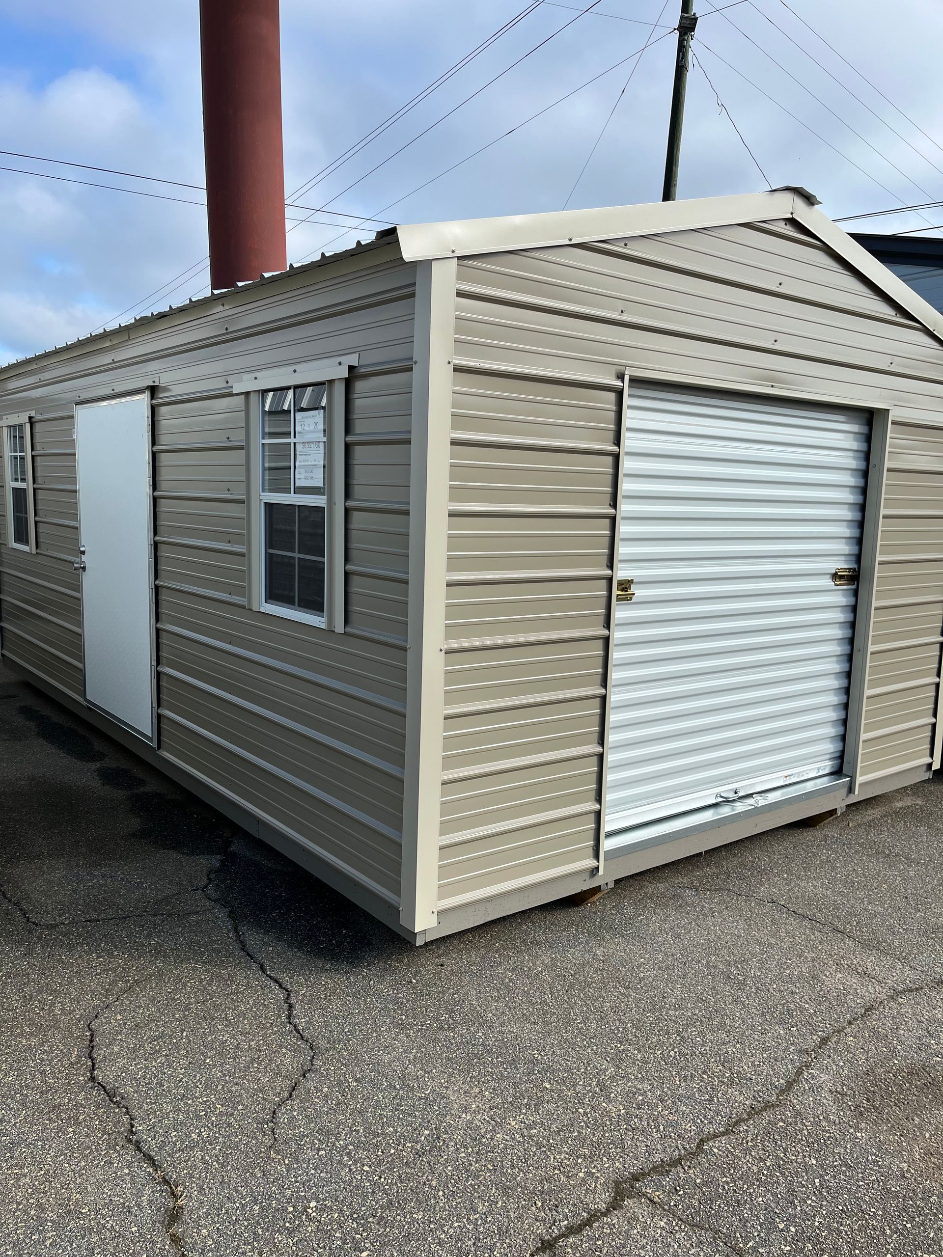 Small beige storage shed with white roll-up door and window, sitting on a paved lot.