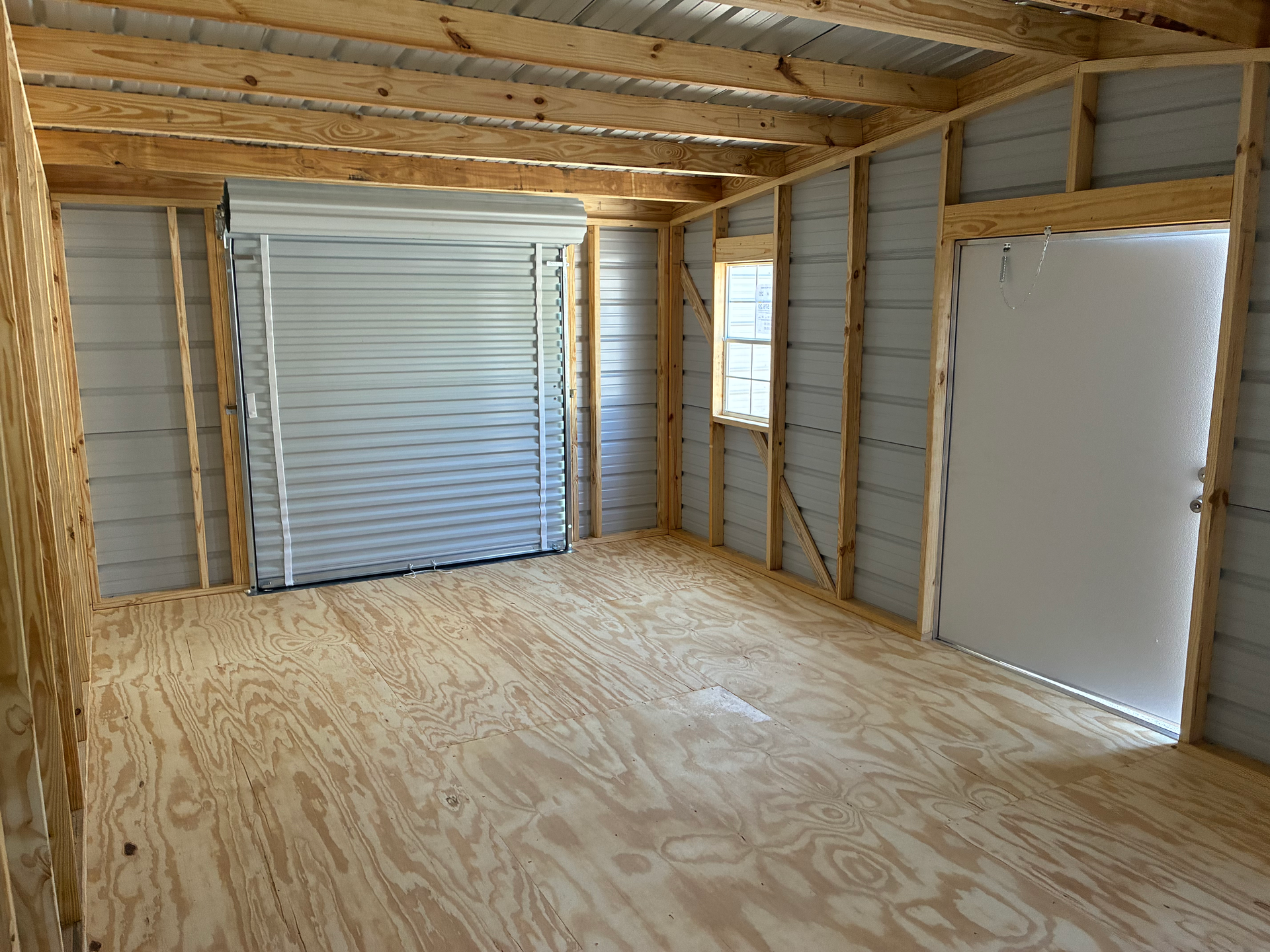 Empty unfinished room with exposed wooden framing, plywood floor, and a white roll-up door and side door.