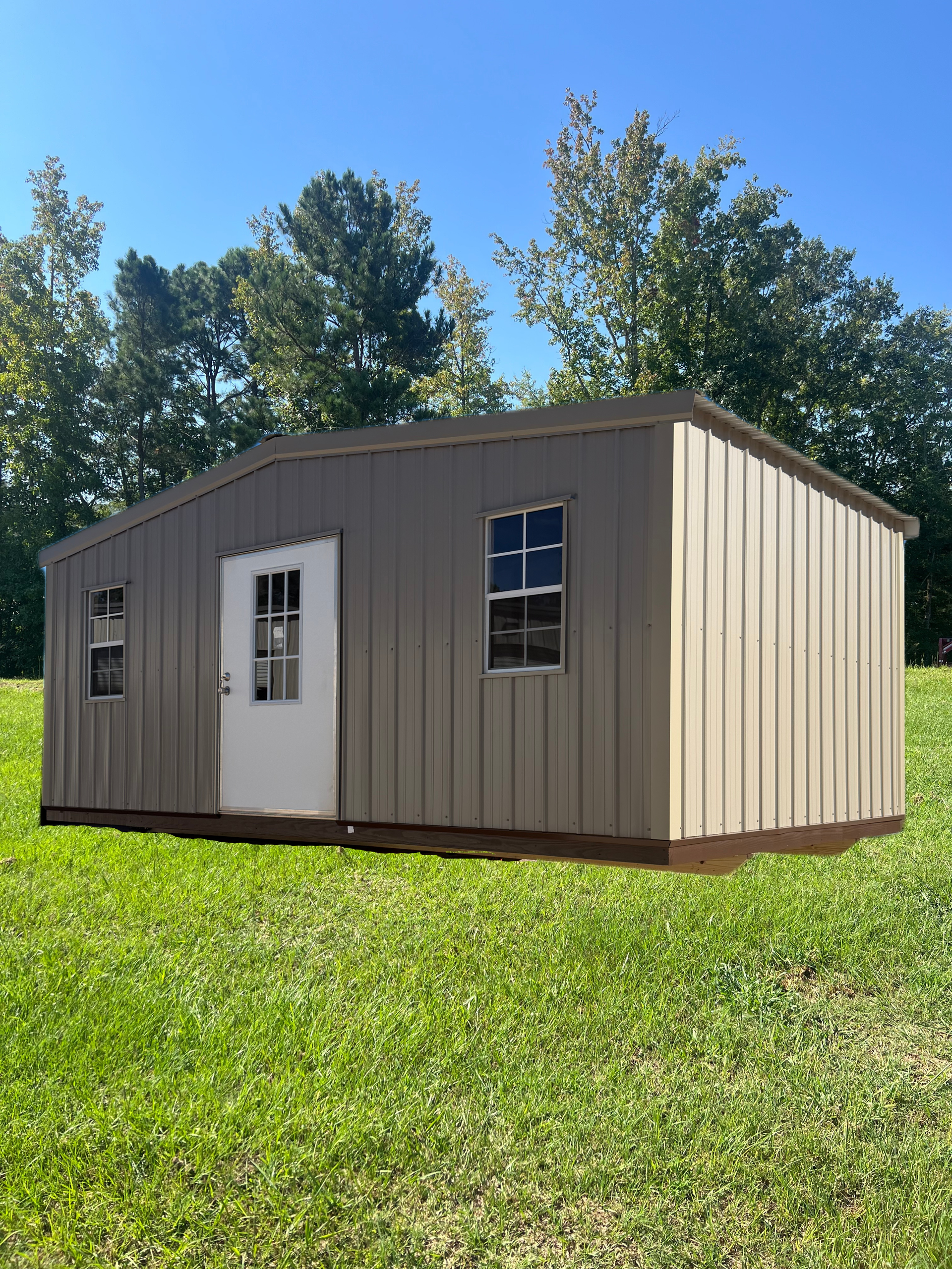 Small beige modular cabin with white door and windows, sitting on a grassy lawn under a clear blue sky.